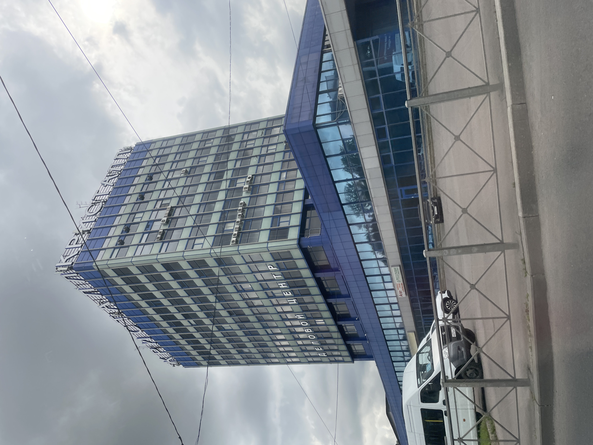 The image shows a tall, blue and white glass building in Saint Petersburg, Russia. The building has "КРИСТАЛЛ" (Kristall) written in large white letters across the top, and "ДЕЛОВОЙ ЦЕНТР" (Business Center) written in smaller white letters on a lower section. The building has many windows and air conditioning units. In front of the building, there is a white van and a dark-colored car parked. A metal fence separates the road from the building's parking area. The sky is cloudy.