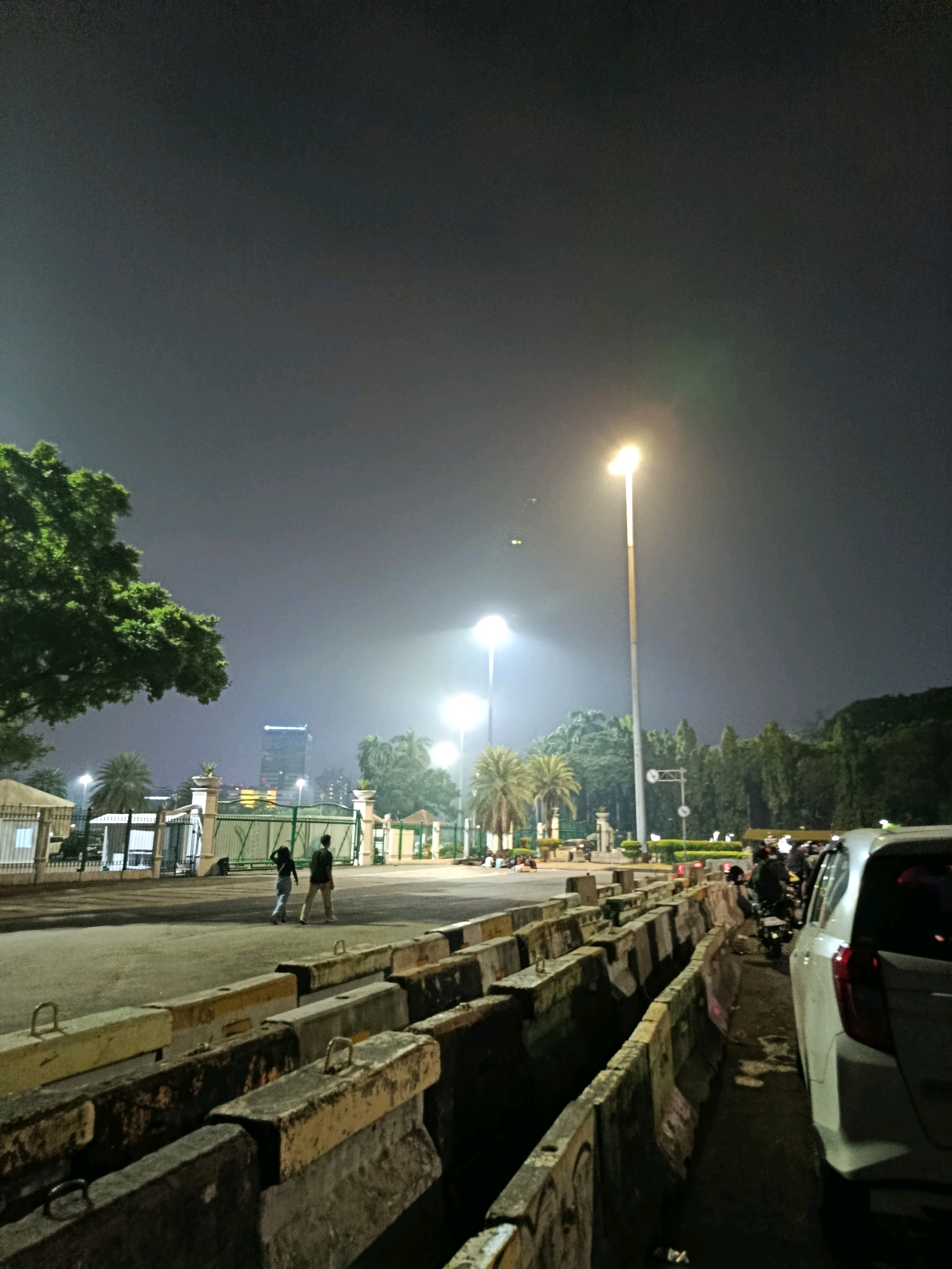 A night scene in Central Jakarta, Indonesia, shows a street lined with concrete barriers and a white car. The sky is dark and hazy, with several tall streetlights illuminating the area. A large tree with green leaves is visible on the left. In the background, there are gates, buildings, and more trees. Two people are walking in the middle distance, and several motorbikes are parked on the right. The atmosphere appears somewhat foggy or misty due to the lighting.