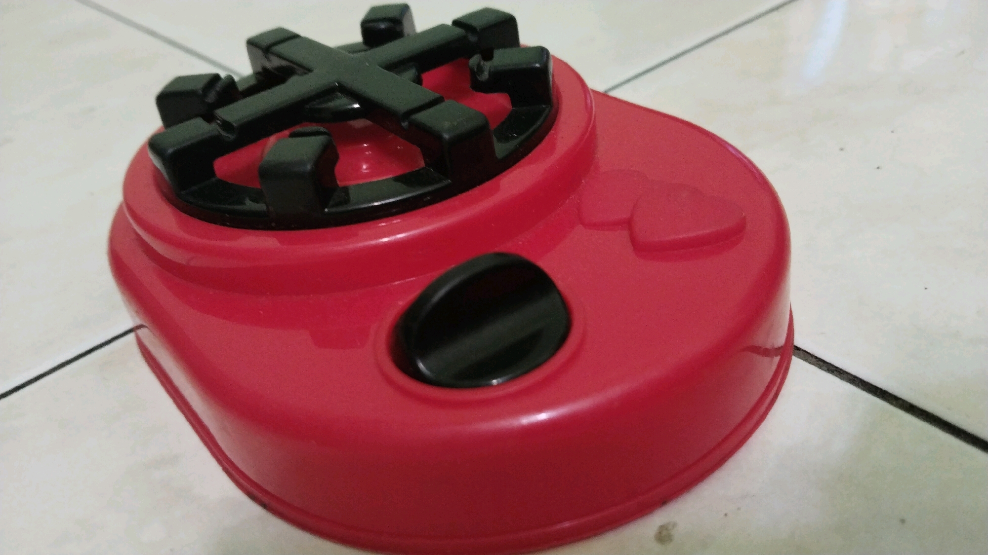 A red toy stove with black stovetop grates and a black dial sits on a tiled floor. The stove has heart-shaped indentations on its side. The scene is set in Simo, Indonesia.