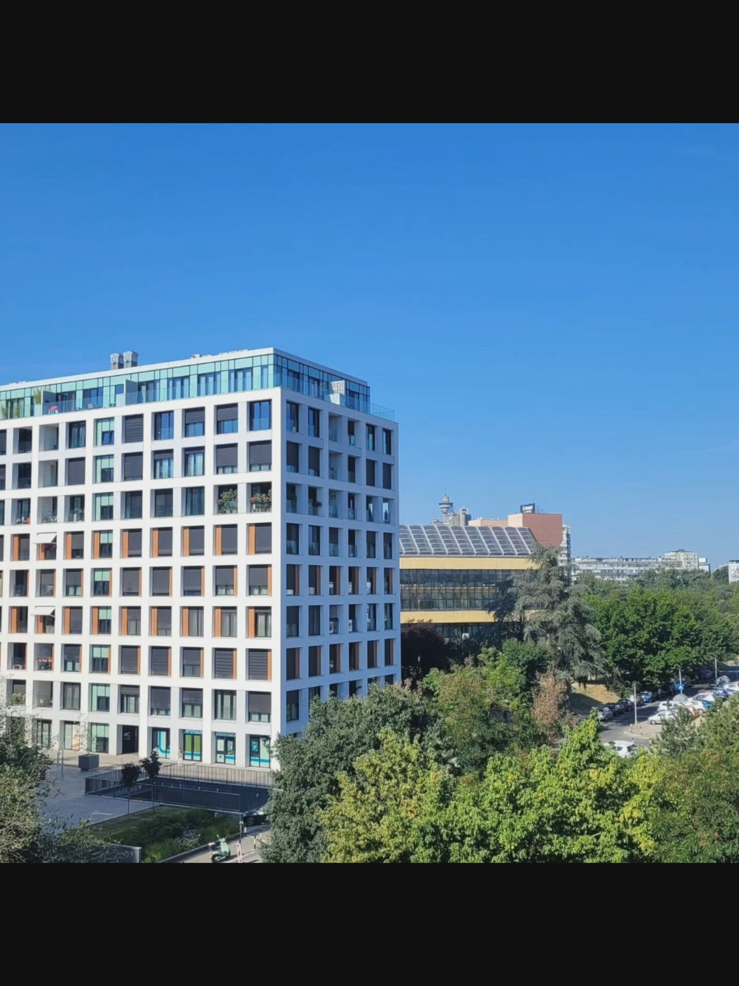 The image depicts a modern apartment building with a white facade and numerous windows, some with closed shutters. The building is situated in an urban environment, with other structures and lush green trees visible in the background. The sky is clear and blue. A street with parked cars and a scooter can be seen in the foreground. The overall scene suggests a bright, sunny day in a developed area of Bielawska Wieś, Poland.