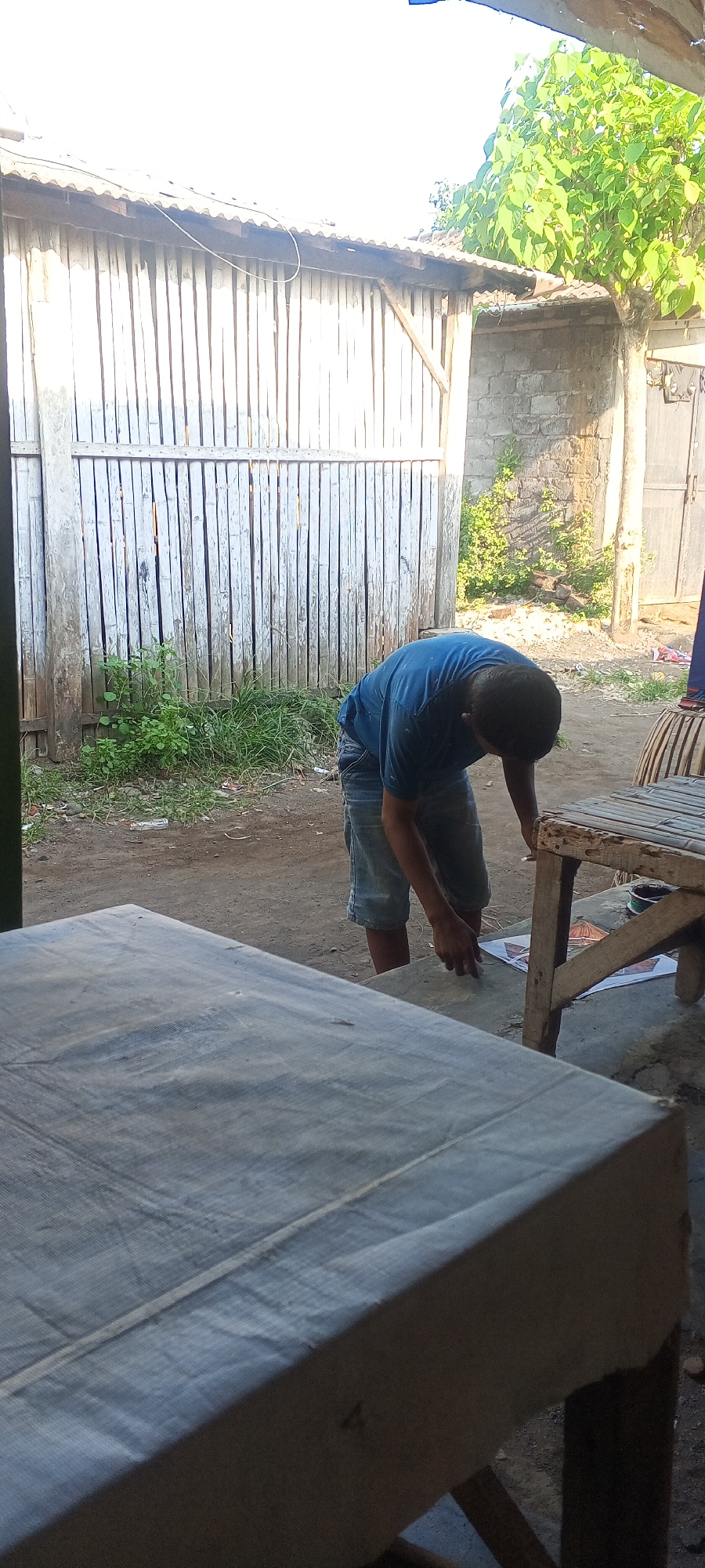 A young man, wearing a blue t-shirt and denim shorts, is hunched over on a dirt ground near a rustic wooden structure. He appears to be working on something on the ground, possibly preparing to fly a kite, as there are kite-like objects visible nearby. Behind him, a weathered wooden fence with vertical planks extends across the background. To the right of the fence, a brick wall and a dense green tree are visible. In the foreground, a large, grey, flat surface, possibly a table, dominates the lower portion of the image. The scene is outdoors, under bright daylight, suggesting it is a casual setting in a residential or open area within Lumajang, Indonesia.