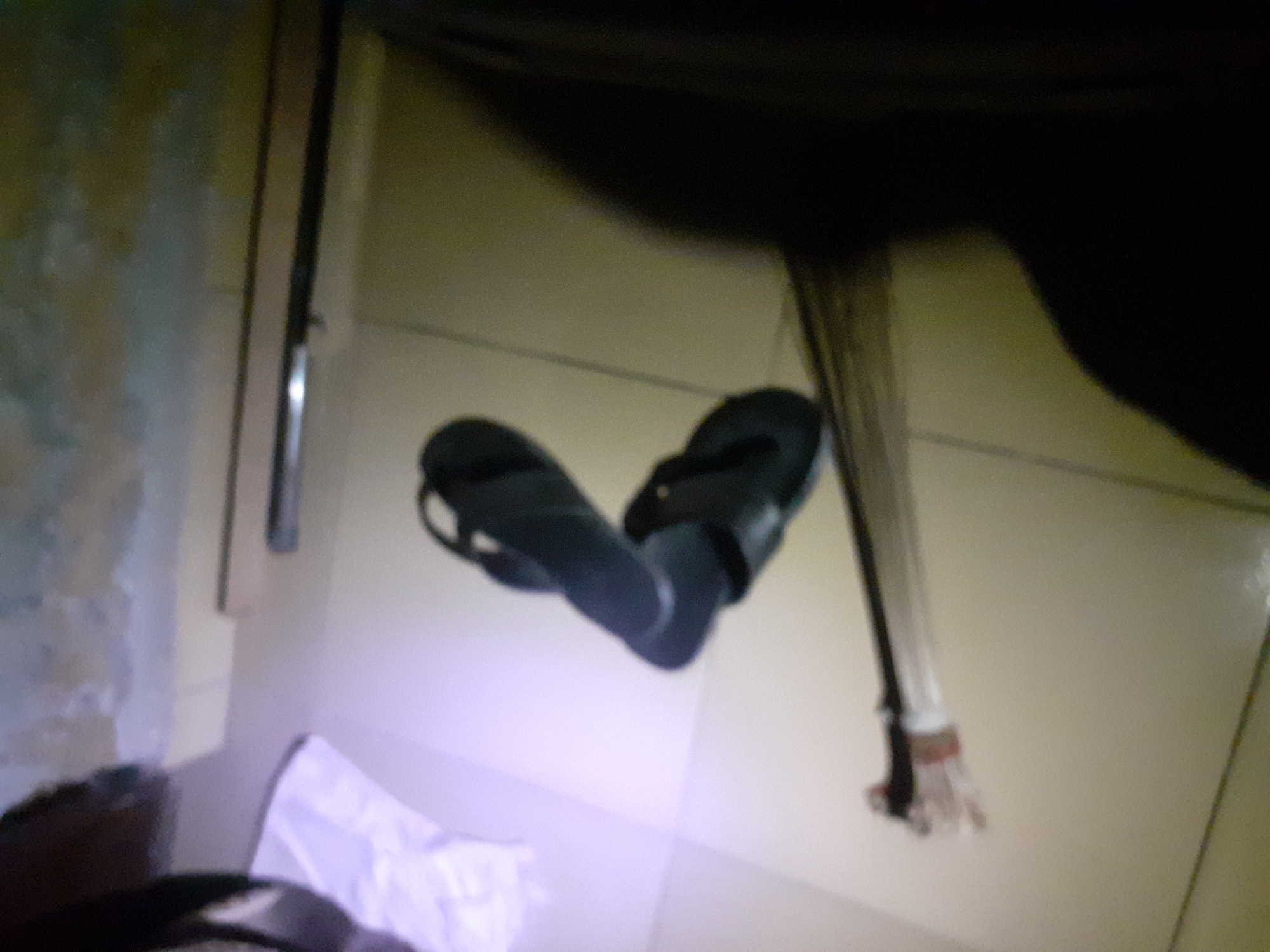 In a dimly lit room, likely in Jalingo, Nigeria, a pair of dark flip-flops are placed on a tiled floor. One flip-flop is positioned slightly ahead of the other, with both appearing to be made of a rubber-like material. A broom with a wooden handle and straw bristles lies horizontally across the floor, intersecting with the flip-flops. The background of the image is blurred, but suggests a wall with some indistinct markings or textures. The overall impression is of a mundane, possibly domestic, scene.