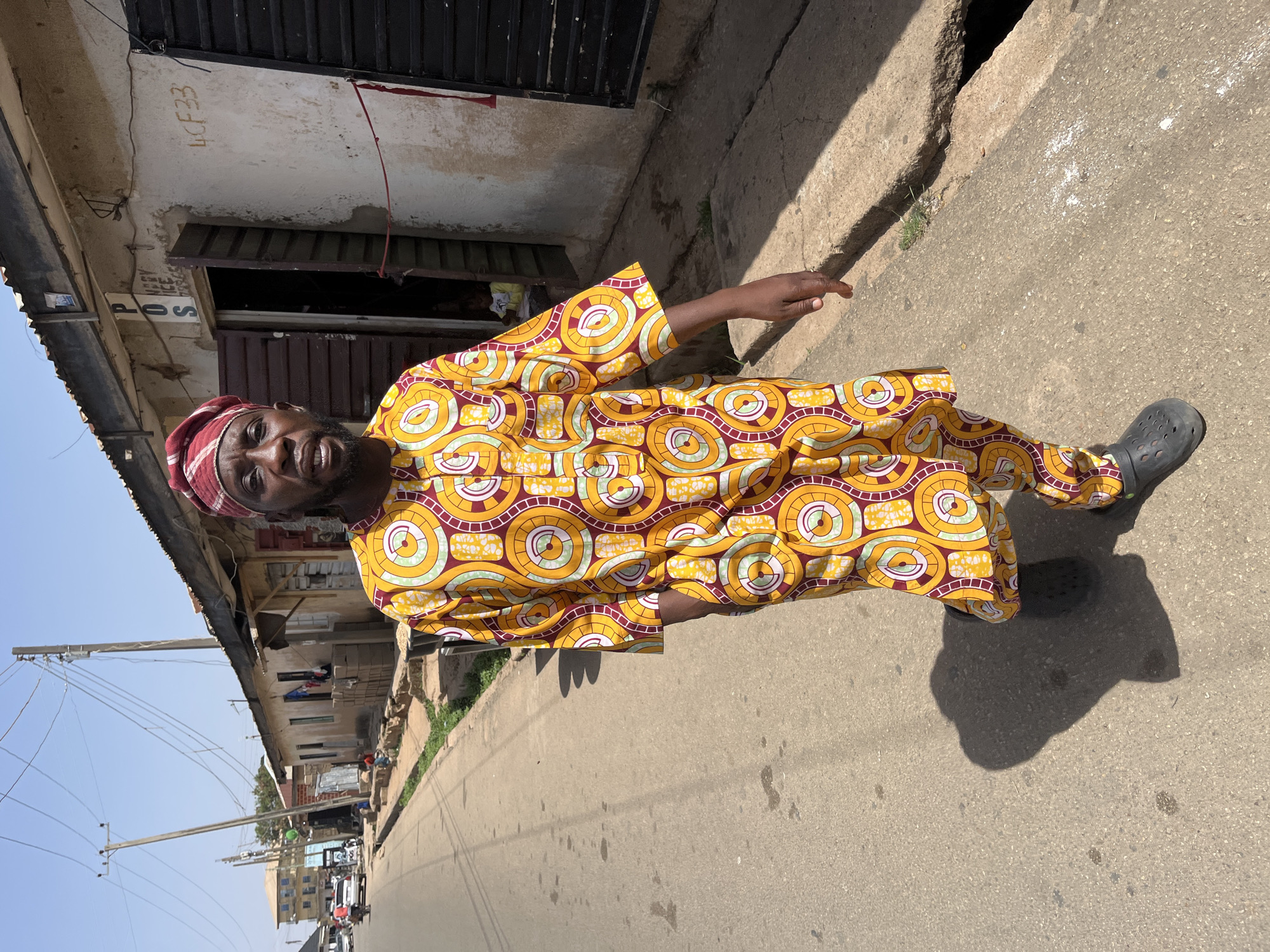 A man stands in a street, wearing a colorful yellow and brown patterned outfit and a red head wrap. He is facing the camera and gesturing with his right hand. He is wearing black shoes. The street appears to be in a densely populated area with buildings on either side, including a building with "LCF33" written on its upper exterior wall. Overhead power lines are visible. In the distance, cars are visible, suggesting a street scene in the city of Jos, Nigeria. The light indicates it is daytime, with a clear sky.
