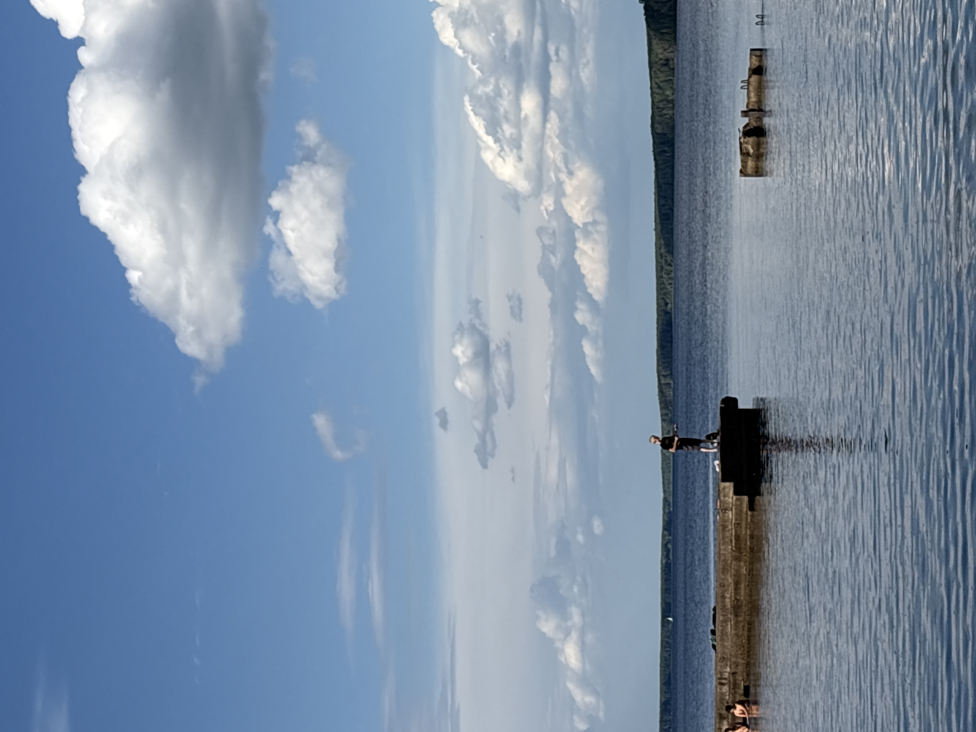 A person is fishing from a concrete pier on a lake, with a line of trees in the distance. The sky is blue with white clouds. Two other people are visible in the water to the left of the pier. A partially submerged structure is in the water to the right of the pier. The scene appears to be the Kama River in Chaikovsky, Russia.