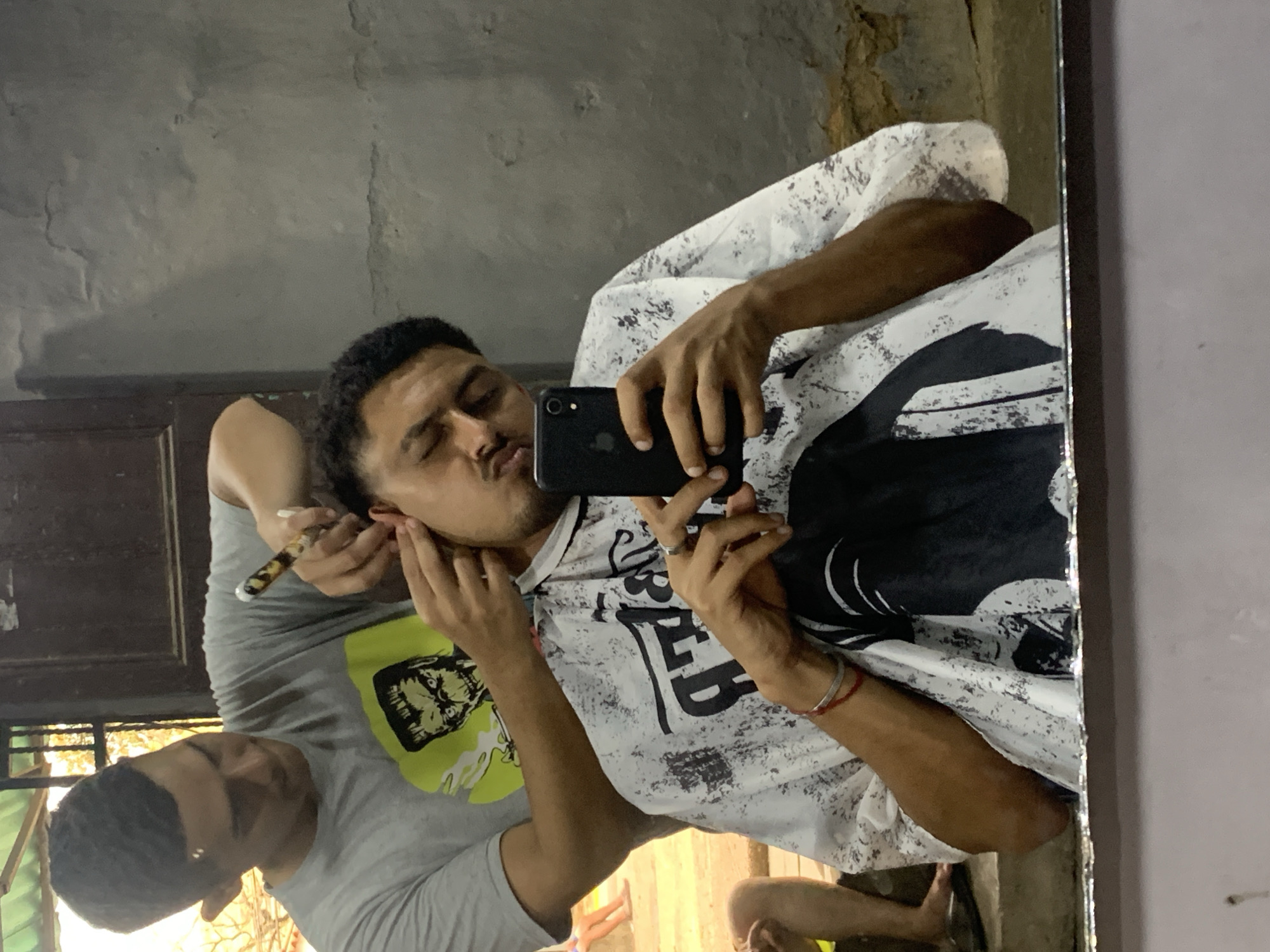 The media file captures a mirror selfie taken by a man who is simultaneously receiving grooming services. The original image is rotated 90 degrees clockwise.
In the center of the reflection, a man with dark hair is holding a black smartphone to take the mirror selfie. He has his eyes closed and lips pursed in a playful expression. He is wearing a white t-shirt with black, abstract patterns and stylized, partially obscured text, which appears to include "OLD" and "BED." A ring is visible on his right hand, and a red string adorns his left wrist.
Partially visible behind the man taking the selfie is another man, presumably a barber or groomer. He has dark hair and is wearing a grey t-shirt with a bright yellow-green graphic print on the front, possibly a stylized face. He is holding a thin, pen-like tool, which appears to be an ear cleaning device or a small razor, near the first man's ear, indicating an ongoing grooming session.
The background reflected in the mirror features a textured, aged-looking grey wall and a dark wooden door or frame. Natural light visible from what appears to be a window or opening in the background suggests it is daytime. The overall setting appears to be an indoor space, possibly a barbershop or a home, characterized by a rustic ambiance in La Tagua, Venezuela.
No other distinct text is clearly legible beyond the partial words on the t-shirt.