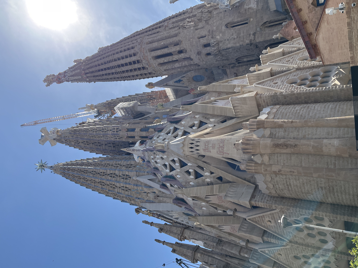 This image captures a low-angle view of the upper sections and intricate details of the Sagrada Familia basilica in Barcelona, Spain. The bright, clear blue sky and the intense sun, partially visible in the upper left, indicate a sunny day, likely mid-day.
The iconic Modernist architecture, characterized by its organic forms, towering spires, and elaborate stone carvings, is prominently featured. A large construction crane is visible behind one of the spires, highlighting the basilica's ongoing construction. Several spires rise towards the sky, one topped with a star and another with a cross. The textured stone facades reveal a wealth of geometric patterns and sculpted elements.
Visible text carved into the stone facades includes the words "Amor" (Spanish/Catalan for "Love"), "Gràcies" (Catalan for "Thanks" or "Graces"), and "Gloria". Other words like "Fides" (Latin for "Faith"), "Deu" (Catalan for "God"), "Jesus", and "Maria" also appear on parts of the basilica, contributing to its profound spiritual narrative. The overall impression is one of majestic scale, intricate artistry, and a continuous work in progress.