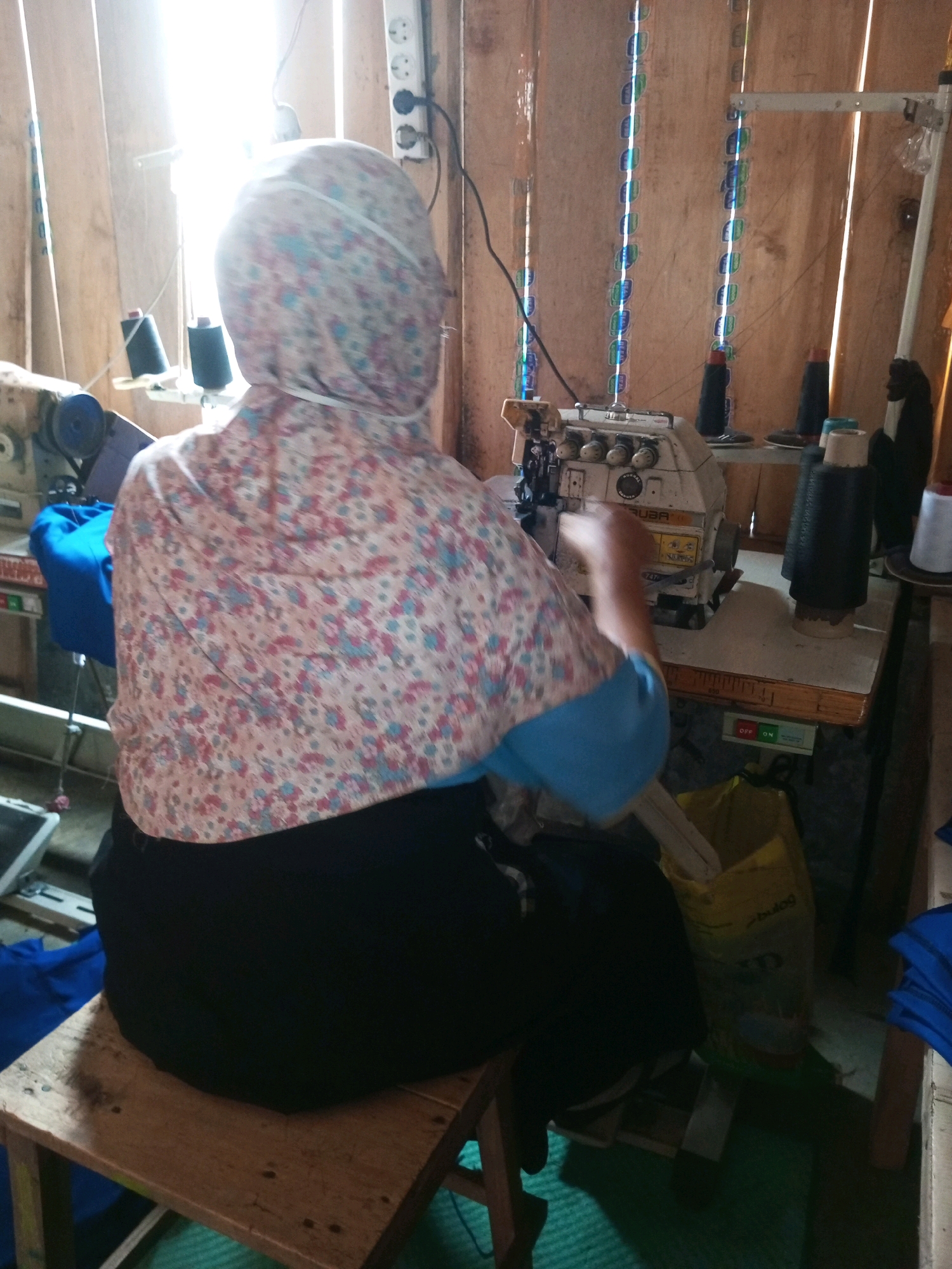 In Kalibeber, Indonesia, a person is seated at a sewing machine in a workshop. The person is wearing a headscarf and a floral-patterned shirt, with a bright blue sleeve visible. They are positioned behind the sewing machine, with their hands presumably operating it. The workshop appears to be rustic, with wooden walls and a bright light source to the left, possibly from a window or overhead lighting, casting strong shadows. Various sewing supplies are visible, including spools of thread in black and white, and a yellow bag filled with materials. To the left of the sewing machine, another sewing machine is partially visible. The overall impression is of a small-scale sewing operation.