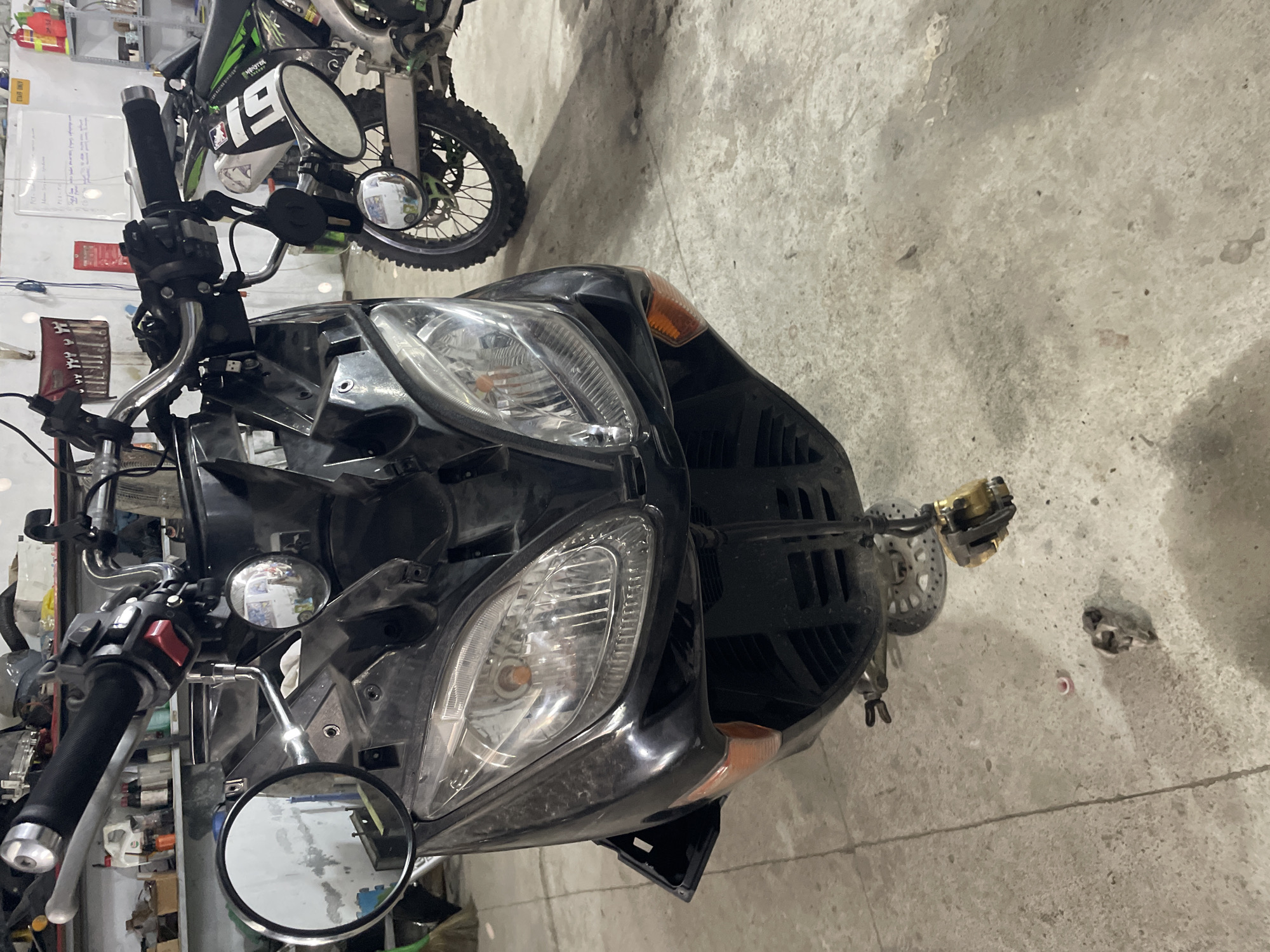 A photograph depicts the front end of a black scooter, seemingly under repair, in a cluttered garage or workshop setting. The scooter's headlight assembly is prominent, with some of its fairings removed, exposing internal components. Handlebars, mirrors, and associated controls are visible. In the background, a green and white dirt bike is partially visible. The floor is concrete and shows signs of use. Based on the prompt, the location is Bac Nha Trang Ward, Vietnam.
