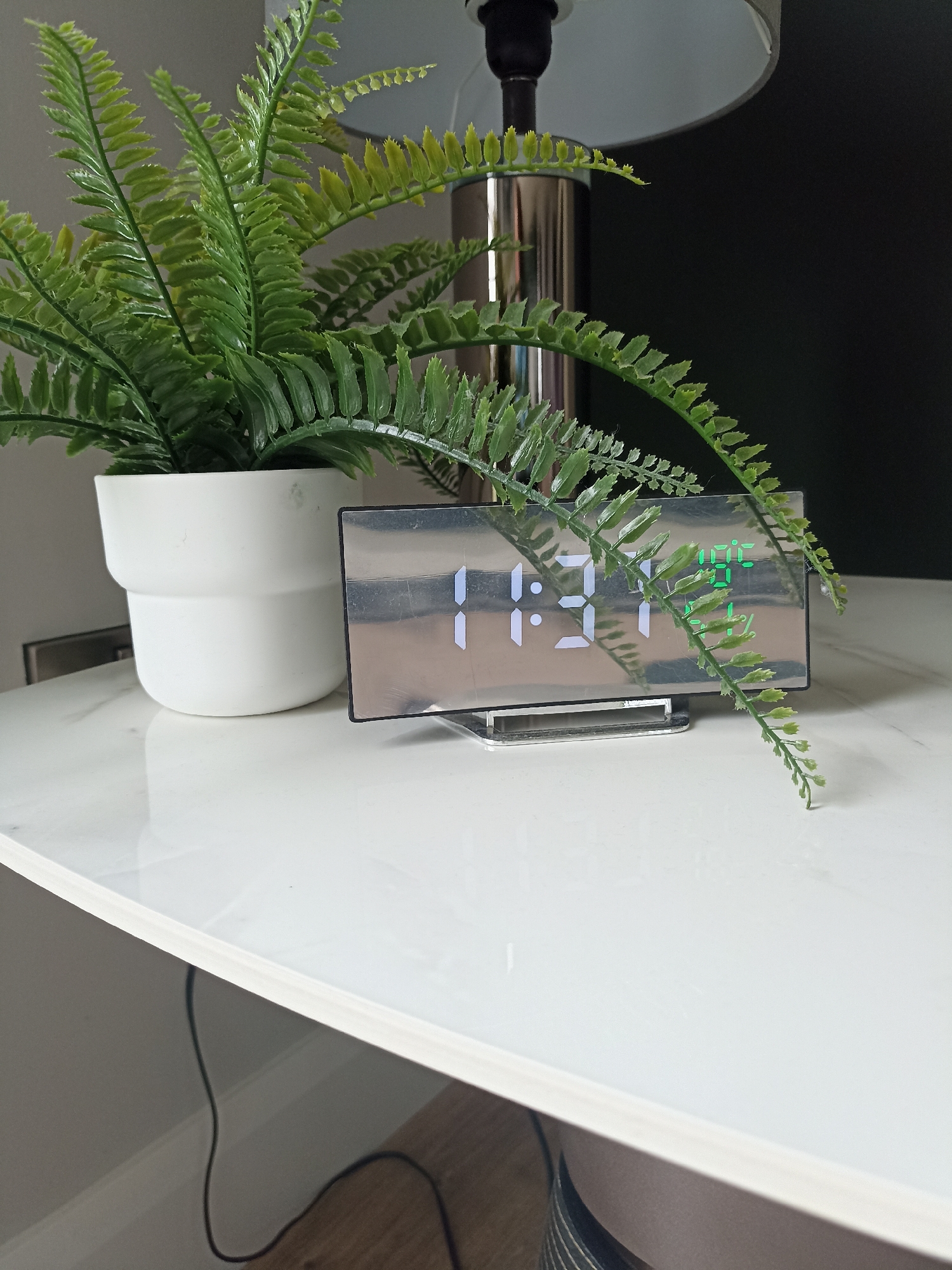 This is a medium shot, eye-level, indoor photograph of a white table with a white ceramic pot containing a fake fern plant, a chrome lamp base, and a digital alarm clock. The alarm clock displays the time 11:21 in white digital numbers, and in the upper right corner, it displays "10:05" in green digital numbers. The clock has a reflective, mirrored surface. The background is a dark wall, and to the left, a portion of a gray wall is visible. A black electrical cord snakes across the lower portion of the image. The photograph does not provide any information about the location in Doirí Beaga, Ireland.