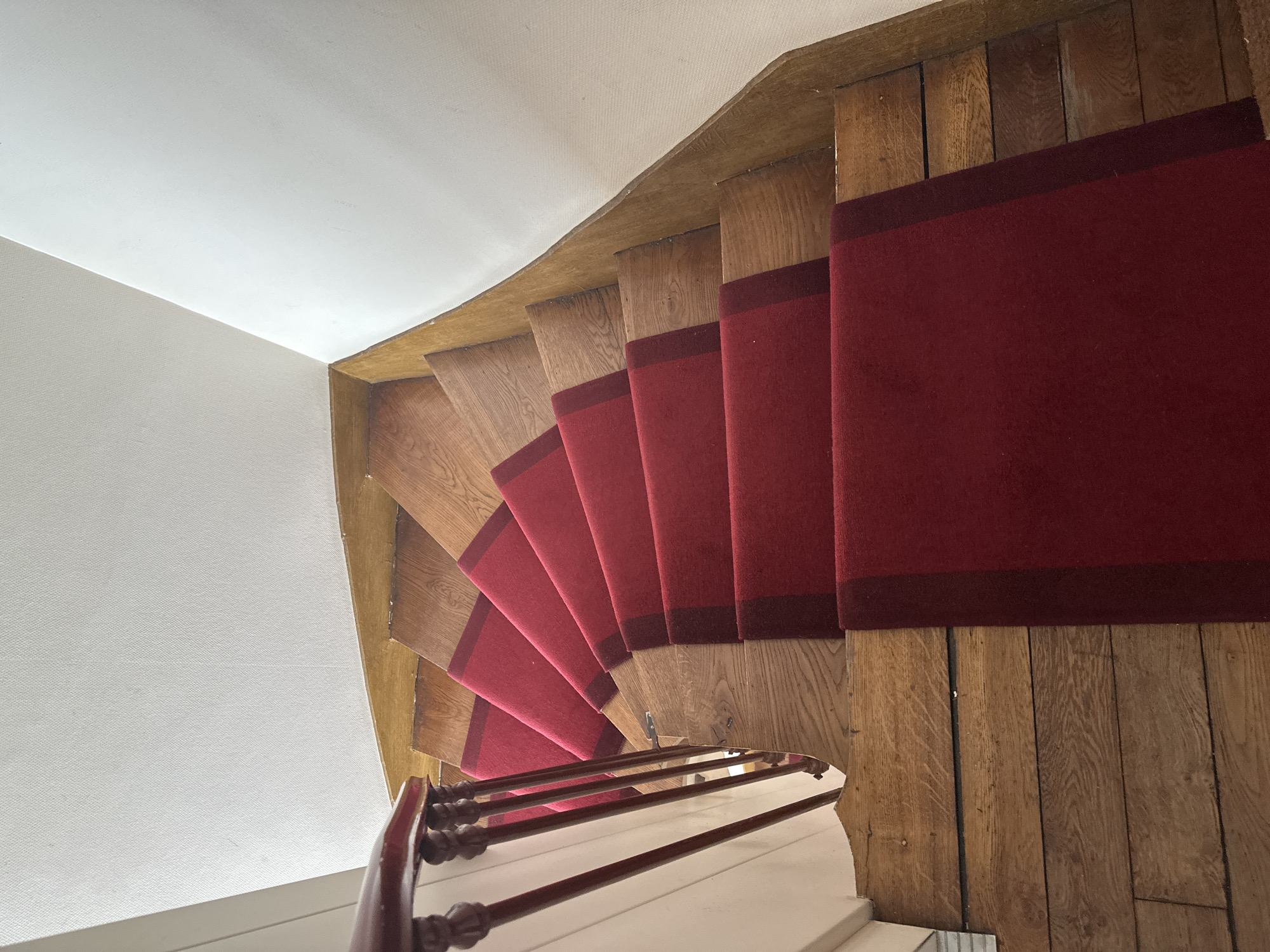The image shows a top-down view of a curved wooden staircase with red carpet runners. The staircase is lined with a dark wooden banister and red-painted railings. The walls are painted white. The scene is indoors and appears to be residential or in a small hotel. There are no people visible in the image. The location in the city of Paris, France, is not specified or deducible from the image.
