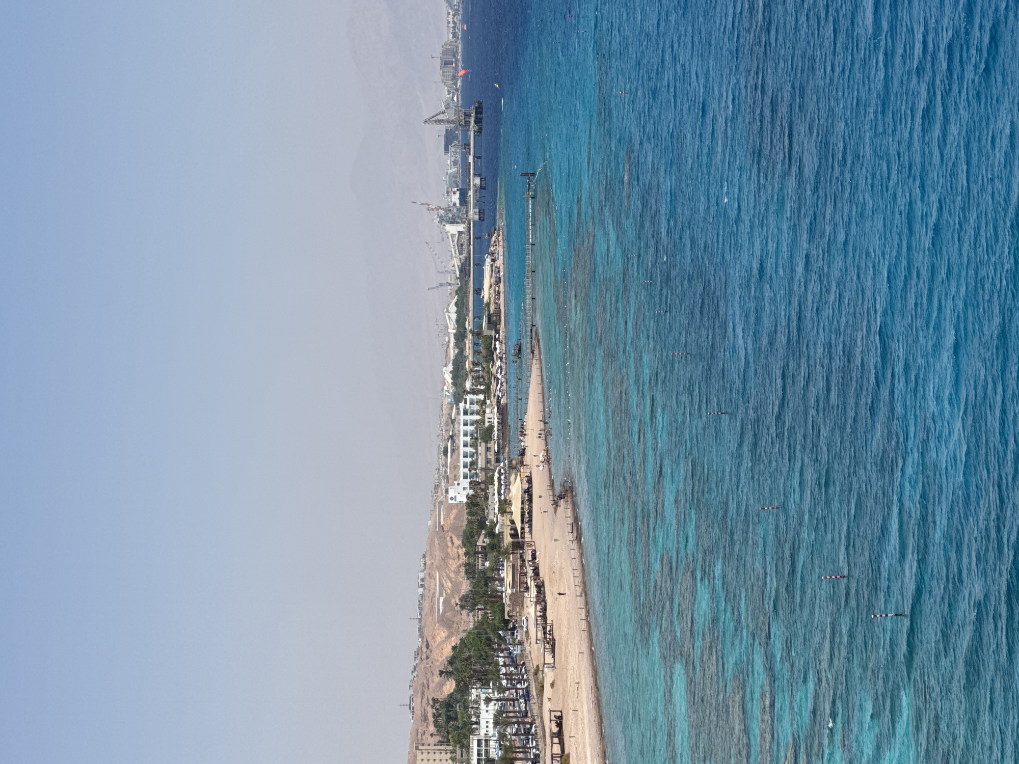 This is a panoramic view of a beach and coastline in Eilat, Israel, likely captured from an elevated perspective. The scene depicts a bright, sunny day with clear blue skies transitioning into hazy mountains in the distance.
In the foreground, the vibrant turquoise waters of the Red Sea are visible, with visible coral reefs just beneath the surface. Several buoys, painted with red and white stripes, are dotted across the water. The coastline features a sandy beach dotted with umbrellas and shaded seating areas, suggesting a popular tourist destination. Palm trees line the edge of the beach, providing shade.
Further inland, a strip of buildings, including hotels and resorts, is visible. A road runs parallel to the beach, with numerous cars parked along it. Beyond the beach and buildings, the landscape rises into arid, hilly terrain with scattered structures and evidence of development, such as roads and construction sites. In the far distance, a harbor or industrial area with cranes and large structures is present.
People are visible on the beach, some walking along the shore, others seated under umbrellas. A few individuals are in the water, swimming or enjoying the sea. The overall impression is of a bustling beach resort area under a clear sky. The specific location within Eilat can be inferred as a developed beach area along the northern Red Sea coast.