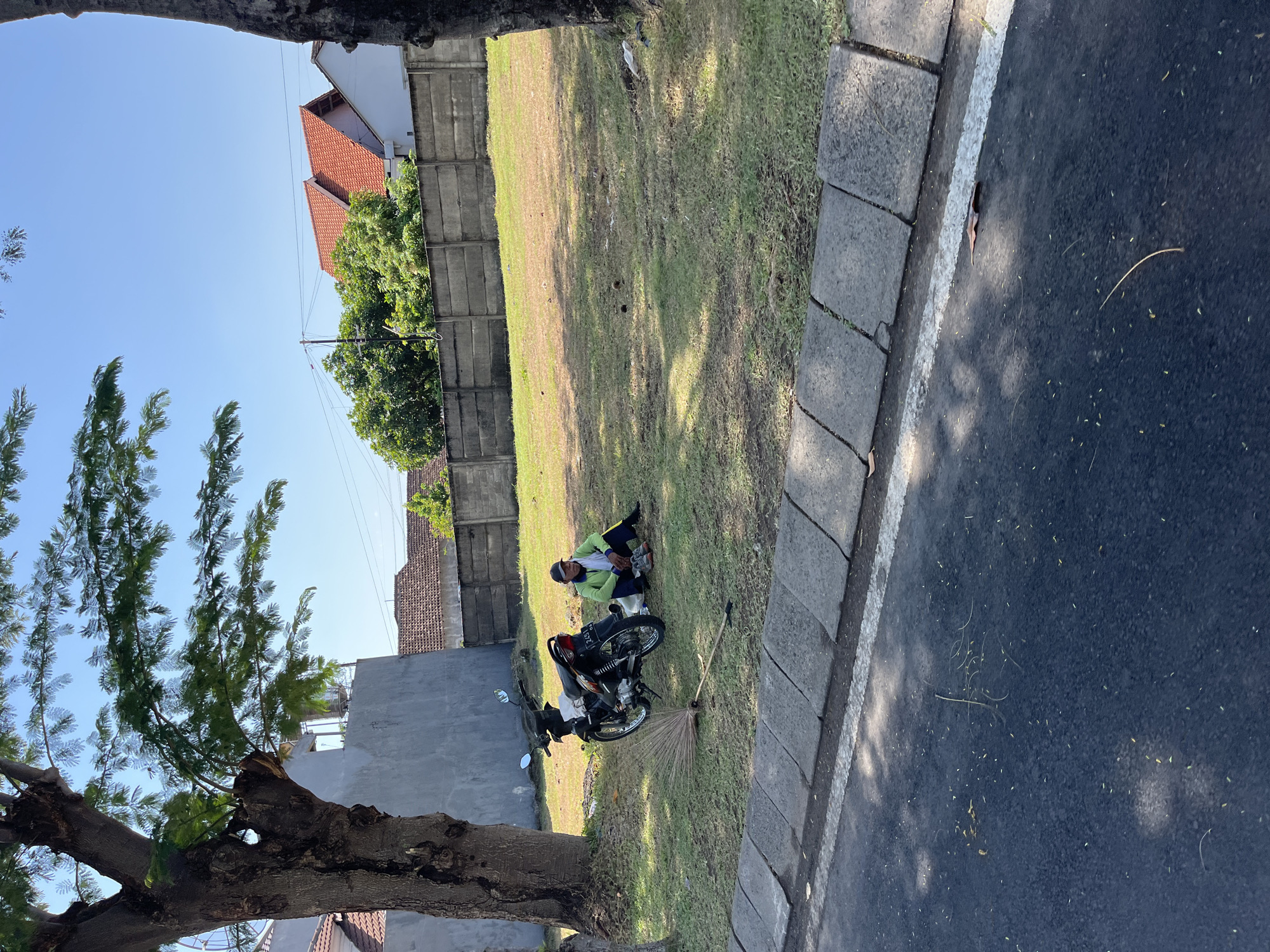 In Entalsewu, Indonesia, a person is seen sitting on the grass near a motorcycle. The person, wearing a green long-sleeved shirt and a dark cap, is positioned to the right of a parked motorcycle. A broom lies on the ground in front of the motorcycle. The scene is set in an outdoor area with a concrete wall and houses in the background, under a clear blue sky. The ground is covered in grass, with trees visible on the left and right sides of the frame. A road with a concrete curb is in the foreground.