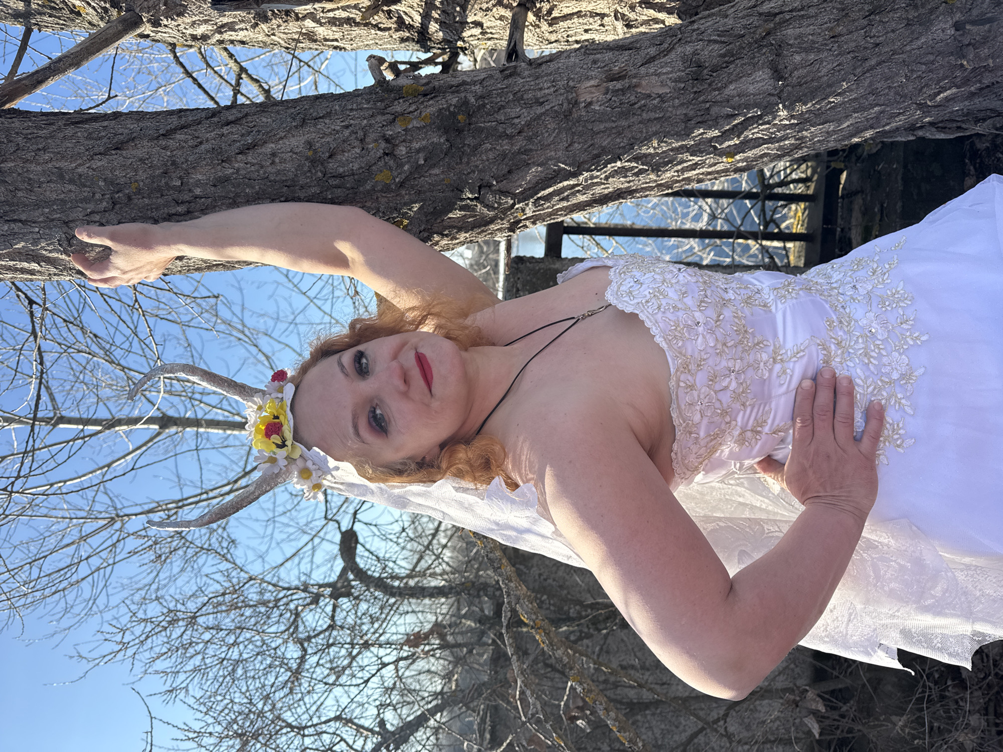 A woman wearing a white dress and horned headwear stands beside a tree branch. The location is identified as the city of Chaikovsky, Russia. The woman is holding onto the branch. In the background is a blue sky and tree branches.