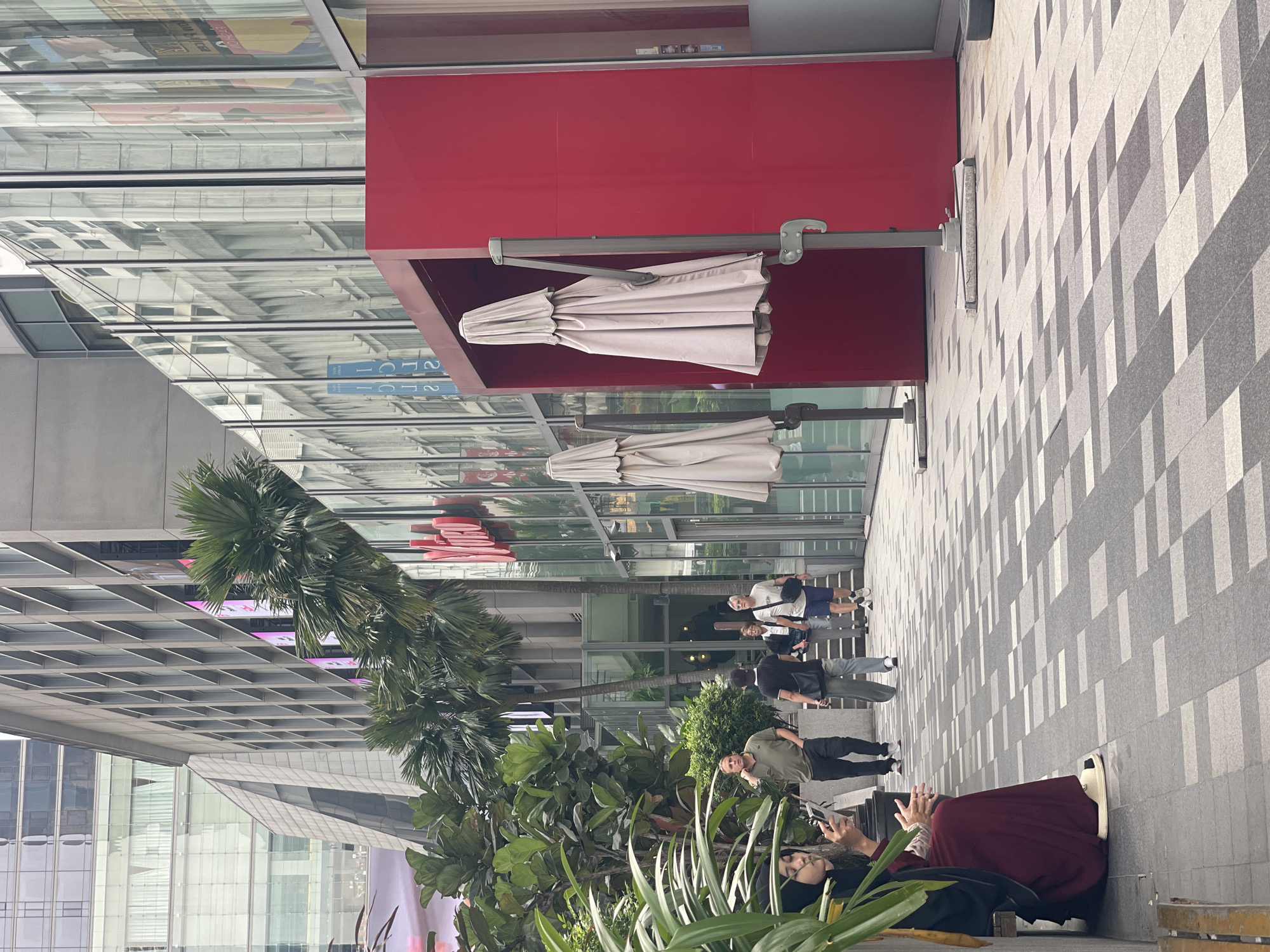 The image depicts an outdoor urban scene, likely in Kuala Lumpur, Malaysia, given the architectural style and the presence of tropical foliage. In the foreground, a woman wearing a dark hijab and a maroon dress is seated on a bench, looking at her phone. Beside her, a man in a green t-shirt and dark pants stands while holding his chin. Further along the paved walkway, which is patterned with grey and white tiles, two more individuals are walking away from the camera. One is wearing a black t-shirt and grey trousers, carrying a black bag over his shoulder. The other is a man in a white t-shirt and blue shorts. The background features a modern building with large glass windows and balconies, interspersed with palm trees and lush green plants. A prominent bright red structure with closed umbrellas in front of it is also a focal point. The overall ambiance suggests a bustling city environment with a mix of people and contemporary architecture.