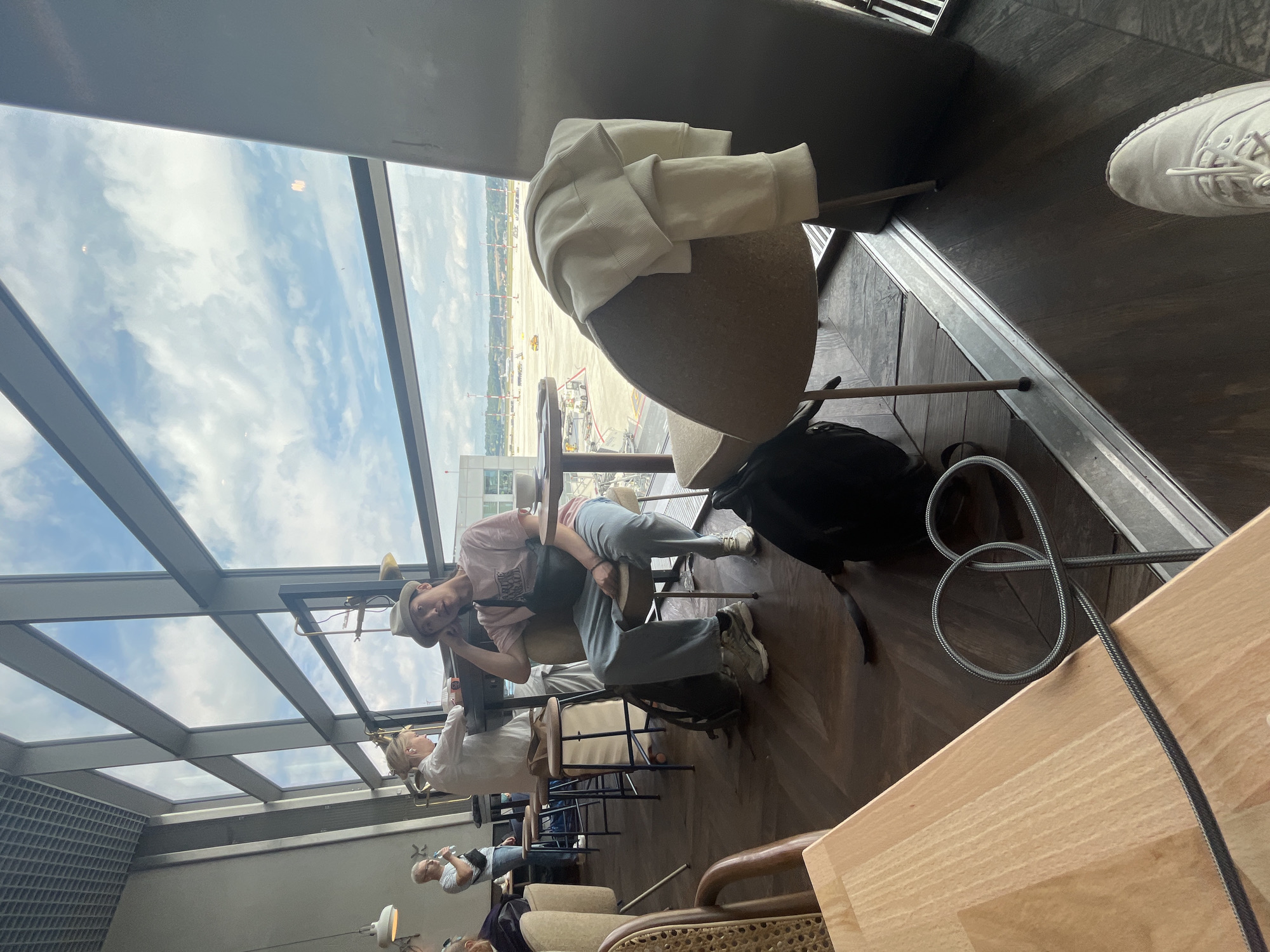 The image depicts a scene inside a modern airport lounge with large windows overlooking an airfield. In the foreground, a young person wearing a hat, a pink t-shirt, and light-colored pants is seated at a table, looking towards the viewer with their hand on their chin. They have a dark bag slung across their body. Behind them, a person in a white shirt is standing at a high counter, and another person in a striped t-shirt is visible further back. A white sweater is draped over the back of a chair in the mid-ground. The flooring is dark wood, and a wooden table is in the lower left of the frame, with a coiled cable on it. The background through the windows shows an airfield with various vehicles, runways, and the sky with scattered clouds. The location is identified as Balice, Poland, suggesting this is likely John Paul II International Airport Kraków–Balice.