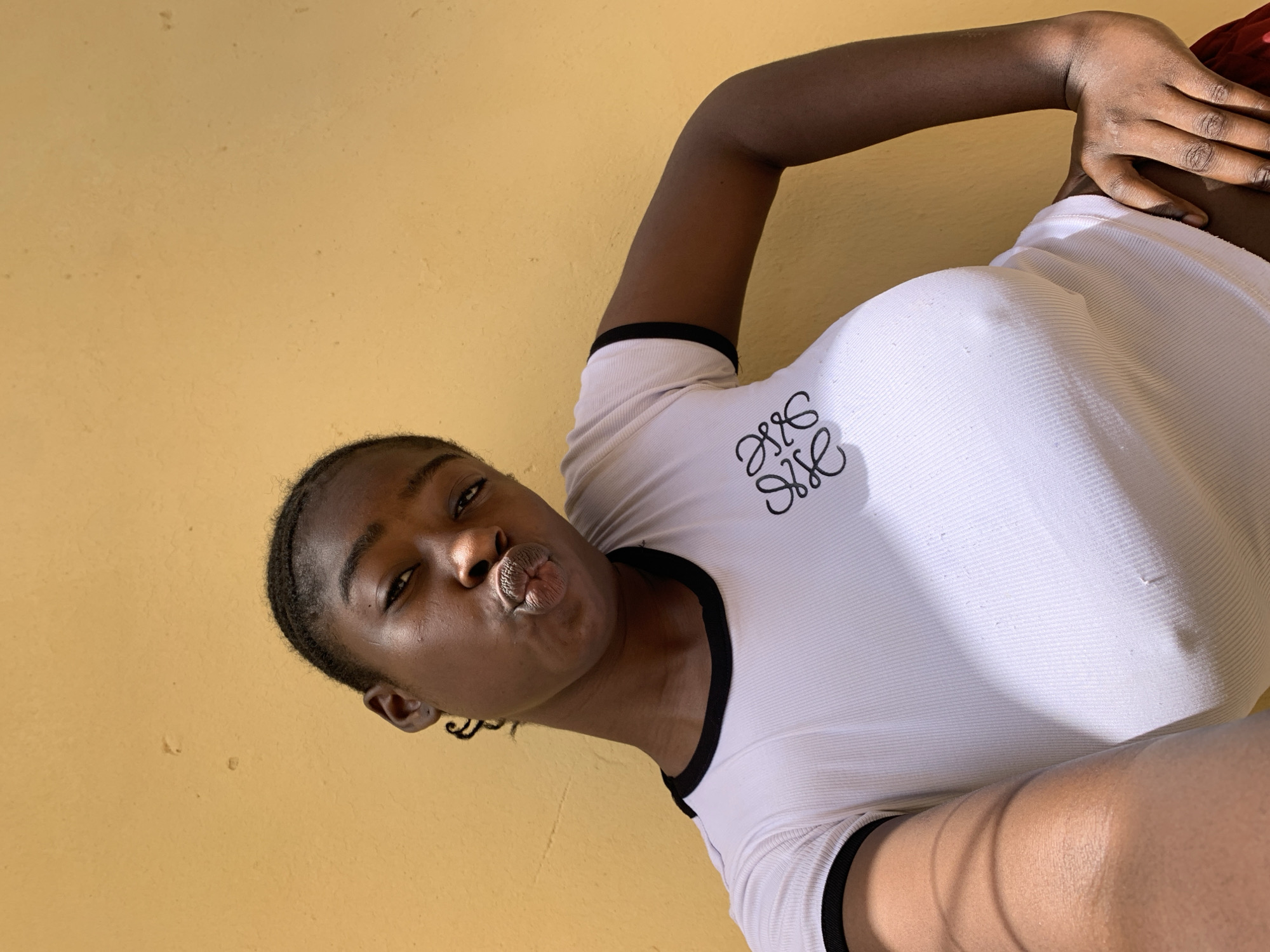 A young dark-skinned woman is posing for a selfie with her lips pursed. She is wearing a white t-shirt with a black trim around the collar and sleeves, and the Loewe logo on the left side. The background is a solid yellow wall. The location cannot be determined from the image.