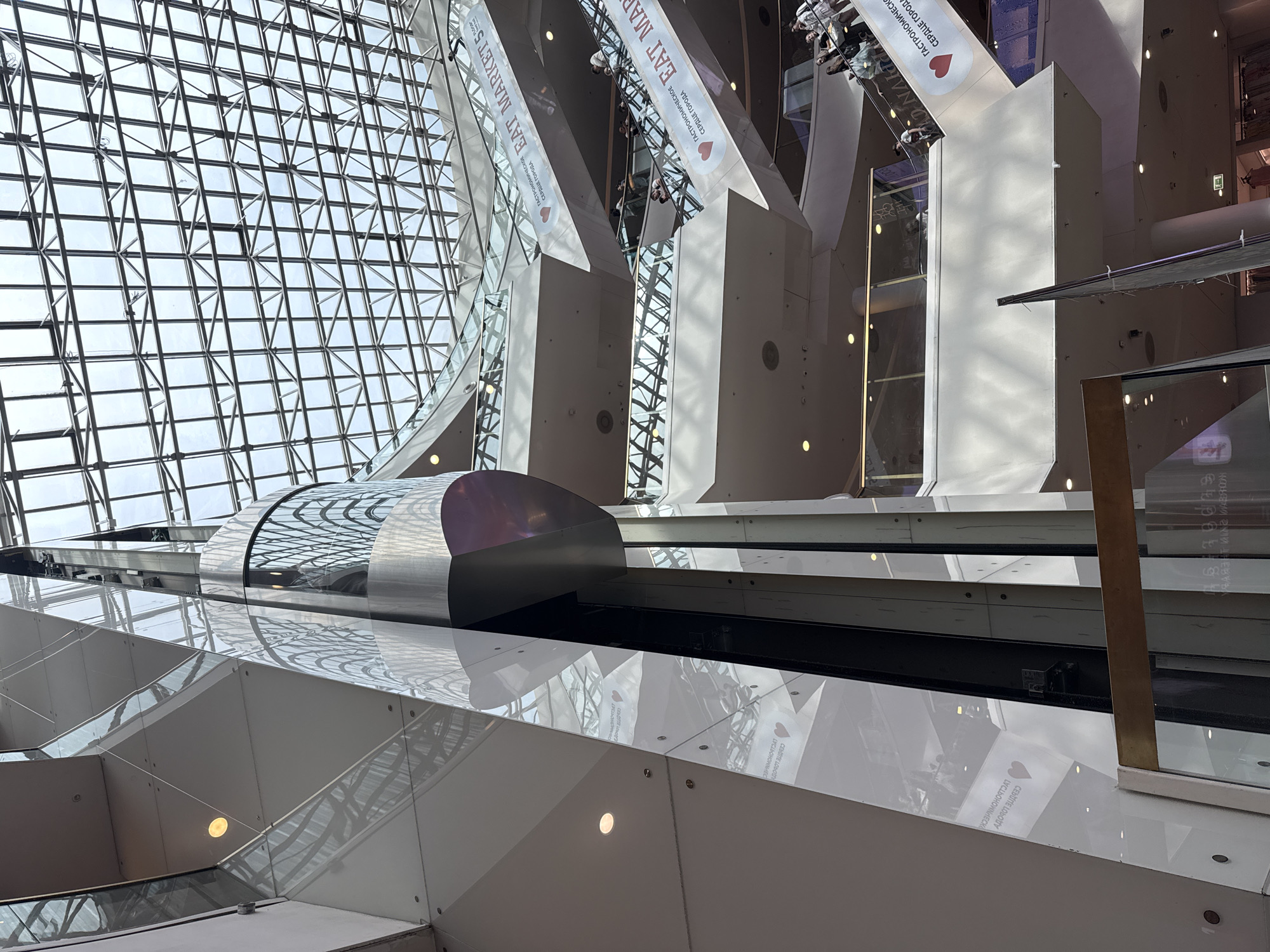 The image captures the interior of a modern, multi-level shopping mall with a distinctive glass and steel architectural design. Dominating the center is a sleek, cylindrical glass elevator shaft that rises vertically through the different floors. The mall features multiple escalators, also with glass enclosures, connecting the various levels. A large, intricate glass and metal grid ceiling allows natural light to flood the space.
Several people are visible on the escalators and walkways, appearing to be shopping or moving between levels. Signs on the escalators display the text "EAT MARKET 5" along with Cyrillic text and a heart graphic, indicating a food market or a section of the mall. The environment is bright and spacious, reflecting the contemporary aesthetic of the building. The reflections on the glass surfaces add to the visual complexity of the scene, showing glimpses of the mall's interior architecture and the people within.