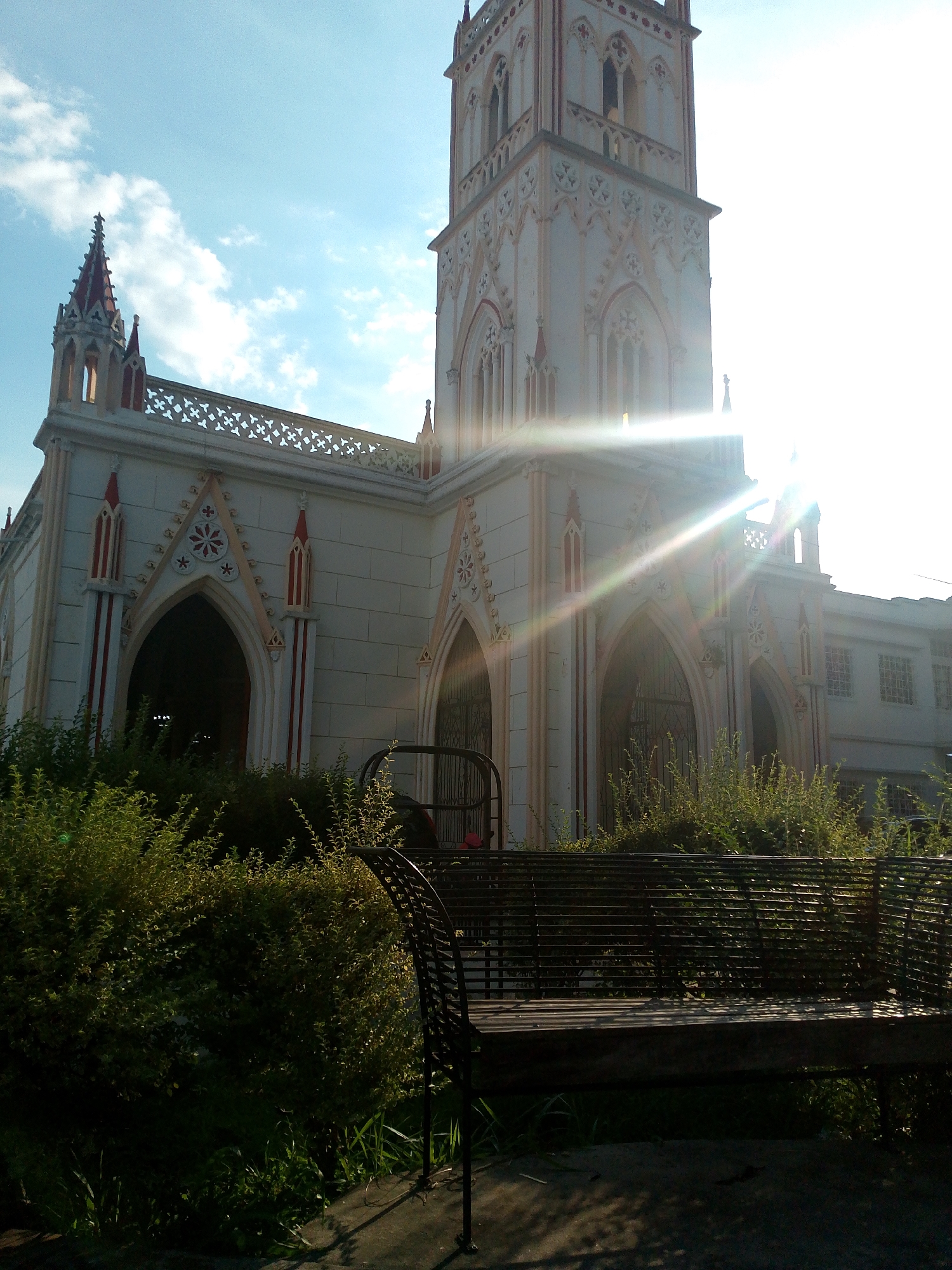 A full shot depicts the Las Lajas Marian Sanctuary in Ipiales, Nariño, Colombia. The church, constructed of light-colored stone with Gothic-style arches and red accents, is partially obscured by overgrown green bushes in the foreground. A dark metal bench is situated in front of the bushes. Sunlight streams over the church, creating lens flare. The sky is light blue with scattered white clouds.