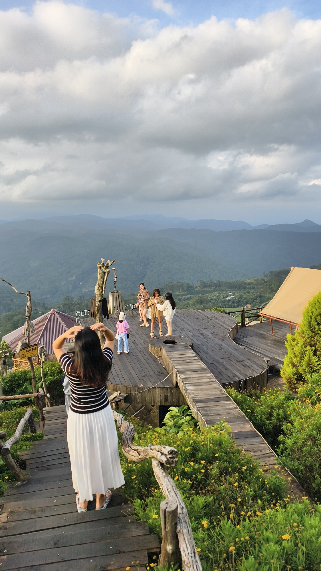 This image captures an outdoor scene at a scenic viewpoint in Phát Chi, Vietnam. In the foreground, a woman with dark hair, wearing a black and white striped top and a white pleated skirt, stands with her back to the camera, adjusting her hair. She is on a wooden pathway that descends down a hillside covered in lush green foliage and small yellow flowers. Further ahead, on a raised wooden platform, four young women are gathered, appearing to be taking photos or posing. The platform is uniquely shaped, resembling a stylized guitar. To the left of the platform, a small building with a purple roof is visible, and a sign with Vietnamese text is attached to a wooden structure. In the background, layers of misty mountains stretch across the horizon under a cloudy sky. A large, tan tent structure is positioned on the right side of the platform, and a dense green tree grows nearby. The overall impression is a tourist attraction offering panoramic views of the surrounding mountainous landscape.