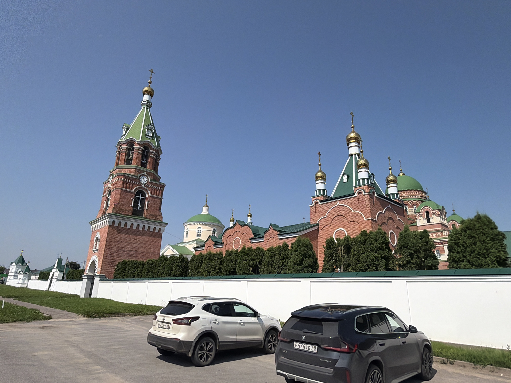 The image depicts a clear, sunny day with a bright blue sky over a religious complex. On the left, a tall, red brick bell tower with a green roof and a golden cross stands against the sky. The tower features a clock face and arched windows. To the right of the bell tower, a red brick church with multiple green domes topped with golden crosses is visible. The church has white decorative elements and arched windows. A white wall runs along the foreground, separating the road from the complex. Two cars are parked on the asphalt road in front of the wall: a white Nissan Qashqai on the left and a dark grey BMW X1 on the right. Green trees and grass surround the buildings and line the wall, suggesting a well-maintained landscape. The overall scene is peaceful and well-lit, characteristic of a daytime view in a Russian town. The location is identified as Troekurovo, Russia.