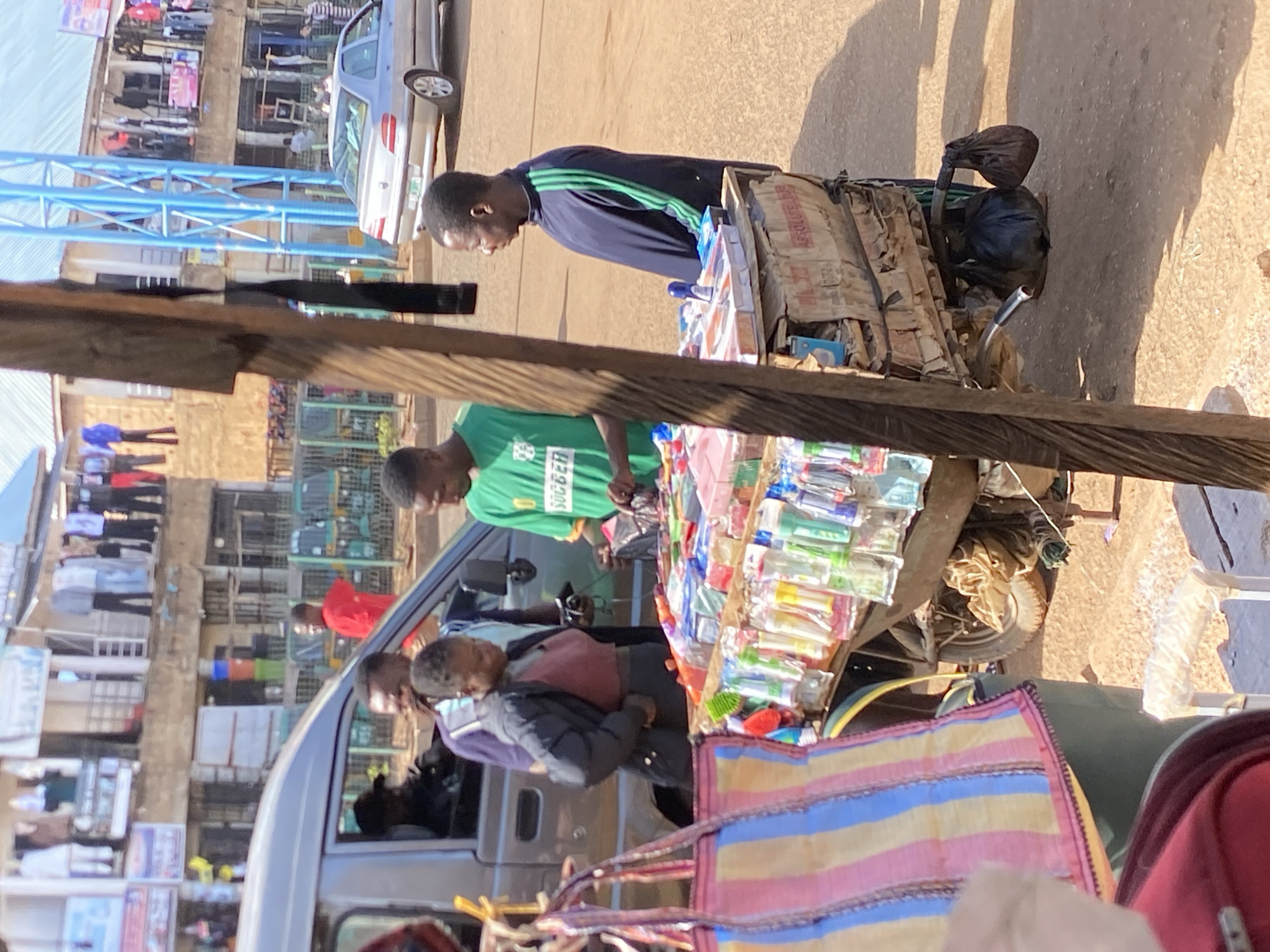 The image captures a bustling outdoor street market scene in Bukuru, Nigeria, under bright daylight. Several individuals are engaged in commercial activities, centered around makeshift stalls.
In the foreground, a man wearing a dark blue tracksuit with green stripes stands by a stall, looking down at the items displayed. Behind a second stall, another man in a green t-shirt with "SUPERGER" or similar text visible is attending to customers. This stall features a wide array of small packaged goods, likely toiletries, snacks, or stationery, neatly arranged on its surface. Another person, possibly a woman, dressed in a maroon top and black jacket, is seen near a light-colored van or minibus, looking towards the stalls.
The scene is set on an unpaved, dusty ground, indicative of a typical market environment. In the background, multiple multi-story buildings line the street, some appearing to house shops or residences, with laundry or other items hanging outside. A blue truss structure, possibly part of a temporary market canopy or construction, is prominent in the middle ground. A silver sedan car, likely a BMW 3 Series, is parked further down the street. Strong shadows suggest it is a sunny day, likely mid-morning or early afternoon. A colorful striped bag, predominantly pink, blue, and yellow, rests in the immediate foreground, partially obscuring the view. Cardboard boxes, one bearing the text "ABSOLUTE," are used as part of the market setup. The overall atmosphere appears busy and functional, characteristic of a local market day.