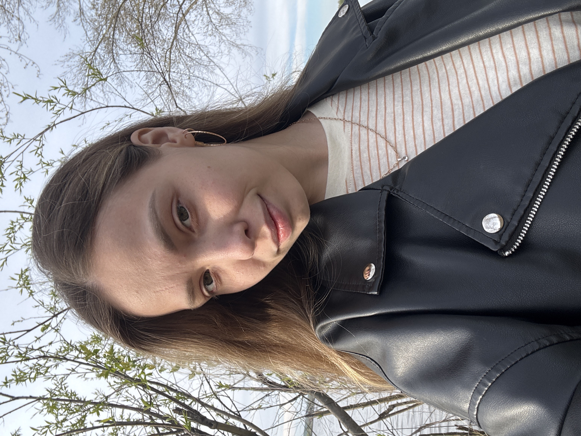 A young woman poses for a selfie outdoors. She wears a black leather jacket over a striped white and peach top, and gold hoop earrings. The background includes bare tree branches and a pale sky. There is insufficient detail to determine the exact location in Chaikovsky, Russia.