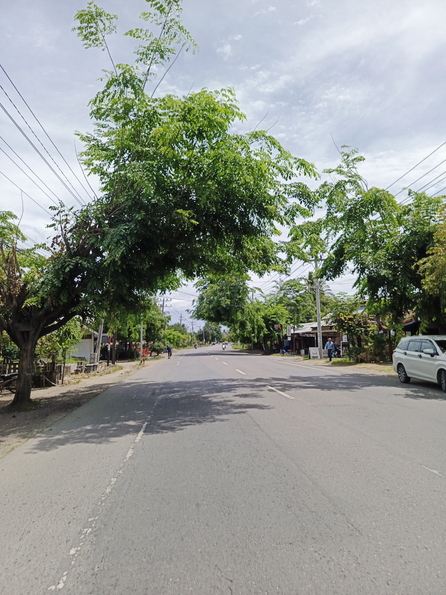 The image depicts a straight, paved road lined with trees and buildings on either side. The road has a white dashed line marking the center. Overhead, power lines are visible, supported by poles. Lush green trees with dense foliage arch over the road, creating a canopy effect in some sections. On the left side of the road, there is a dirt shoulder with a tree and some vegetation. Further down the road on the left, several small buildings are visible, some with signs. On the right side of the road, there are more buildings, a white van, and a person walking. A few motorcycles and a person on a motorcycle are also present on the road in the distance. The sky is partly cloudy with a bright, hazy atmosphere. The scene appears to be a typical street in a populated area, possibly a village or small town, in Cotbadatunong, Indonesia.