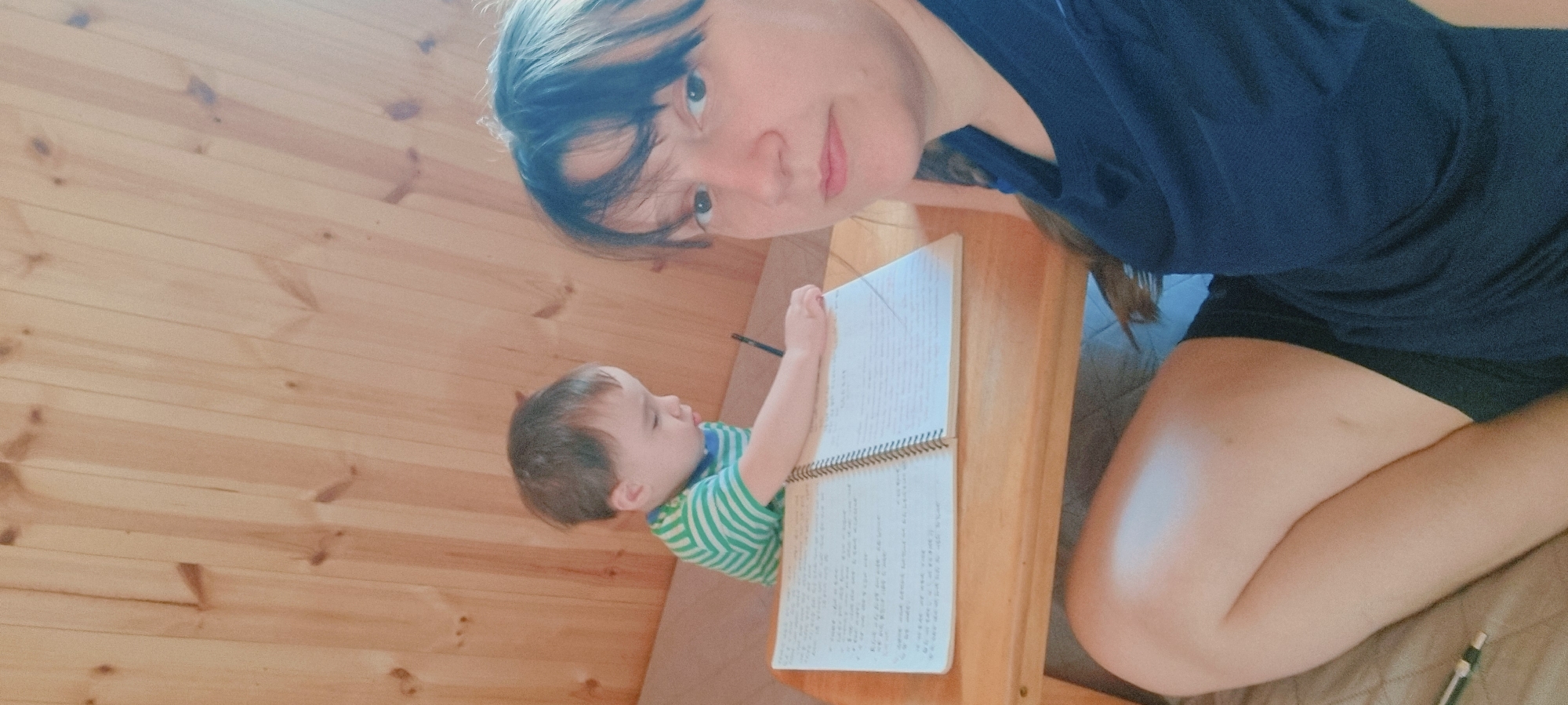 A woman and a child are near a table. The woman is wearing a blue shirt and black shorts. The child is wearing a green and white striped shirt. The child has a writing utensil in their hand. The table has an open spiral-bound notebook on it. The interior walls are made of light-colored wood. The location is inferred to be in Dunnae-myeon, South Korea.