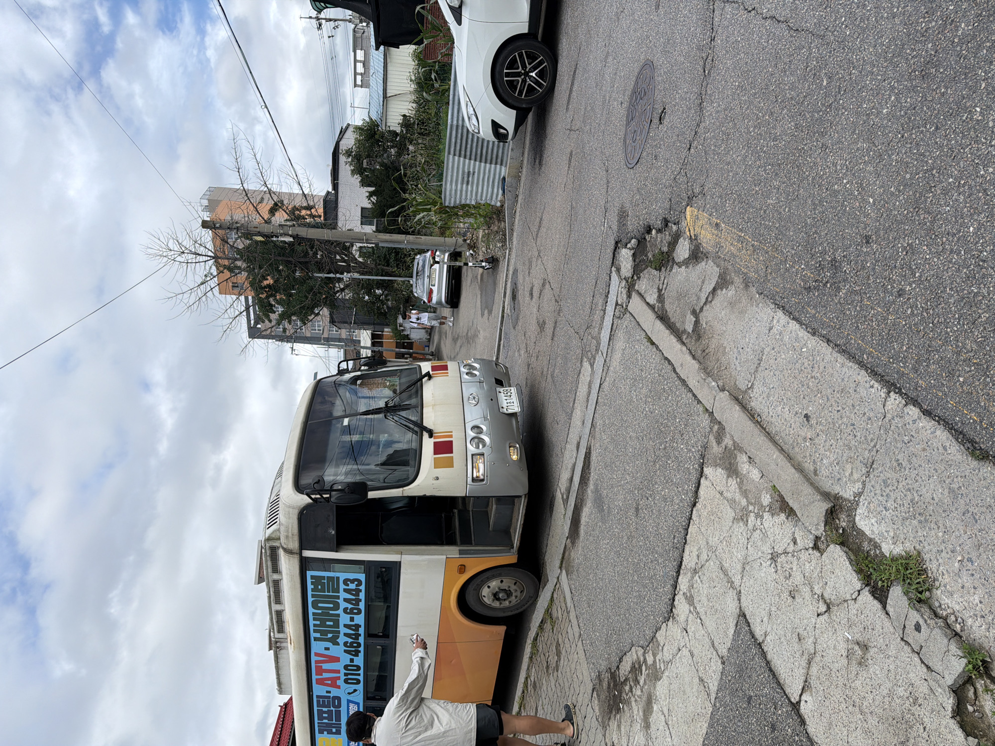 In Chuncheon-si, South Korea, on a cloudy day, a white and orange bus is parked on the side of a narrow asphalt road. The bus has Korean text and a phone number, "010-4644-6443," visible on its side. A man wearing a light-colored long-sleeved shirt and dark shorts stands near the bus, holding what appears to be a small white object. He is facing away from the bus, towards a white car parked further down the street. The road is cracked and shows signs of wear. In the background, there are buildings, a bare tree, and overhead electrical wires. A white SUV is parked on the right side of the road, partially obscuring a corrugated metal fence.