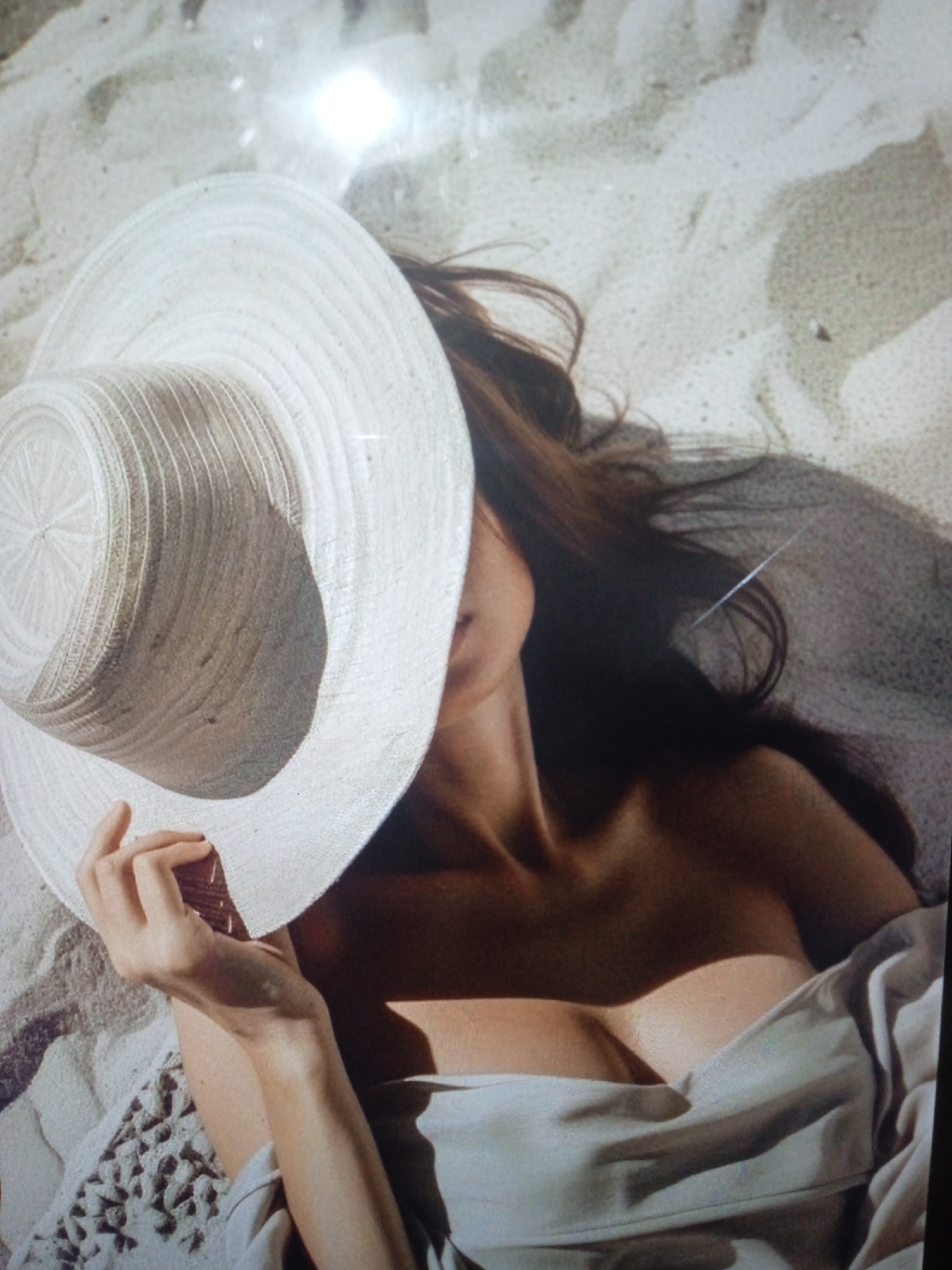 A person lies on a sandy surface, partially obscured by a large, white, wide-brimmed hat. Their hair, dark brown and flowing, spreads out on the sand. The person is wearing a light-colored garment that drapes around their upper body, revealing a portion of their chest. Their left hand rests on the brim of the hat. The environment appears to be a beach or a sandy area, with dappled light and shadows suggesting an outdoor setting, possibly during daytime. There is no discernible connection to the city of Chernivtsi, Ukraine, as the image lacks any specific geographical markers or context related to that location.