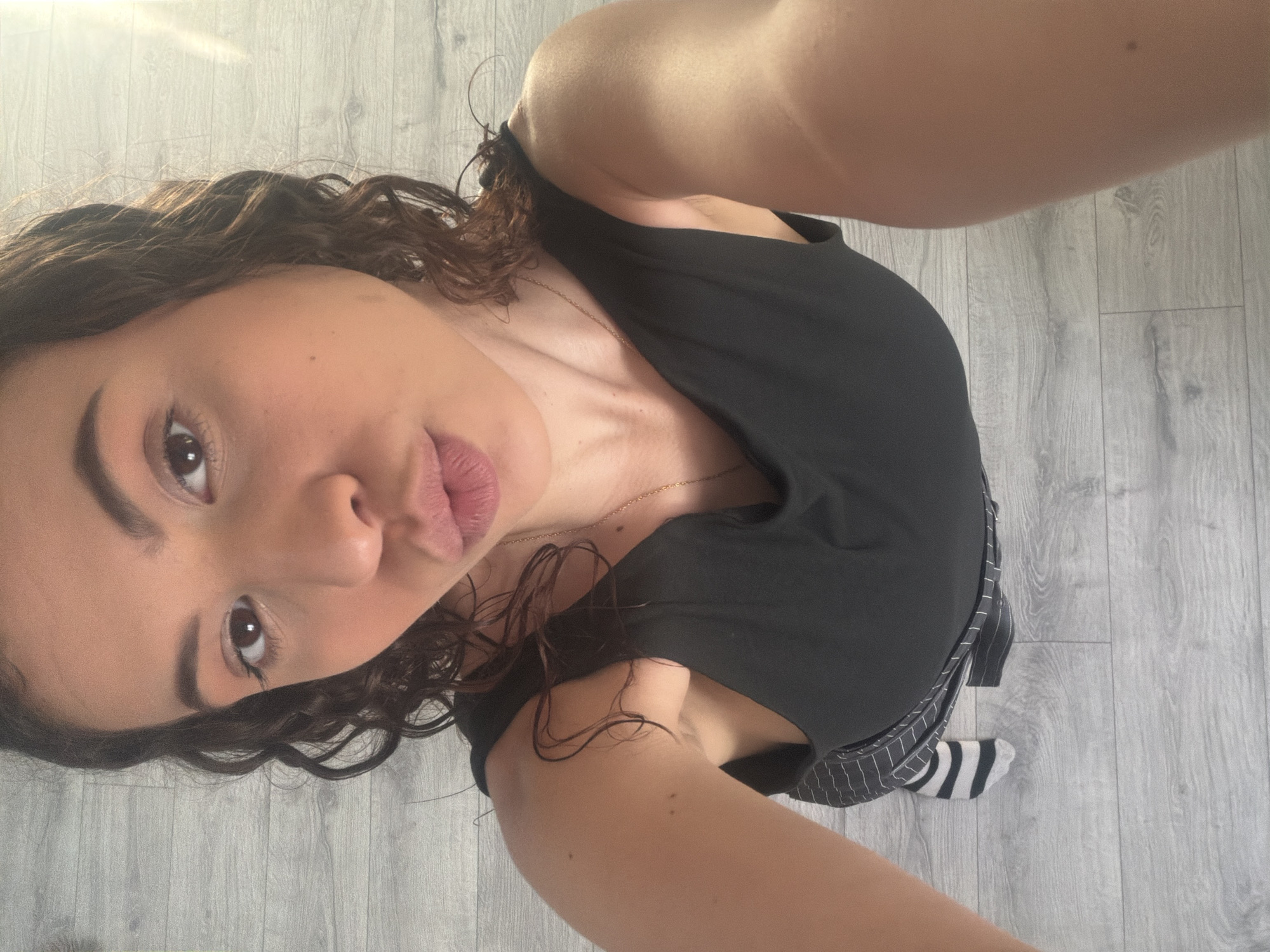 A high-angle, close-up, selfie-style shot shows a young woman with dark curly hair and fair skin, puckering her lips. She is wearing a black tank top and a striped skirt. The background is a wooden floor. The lighting suggests an indoor setting. There are no visible signs of the city.