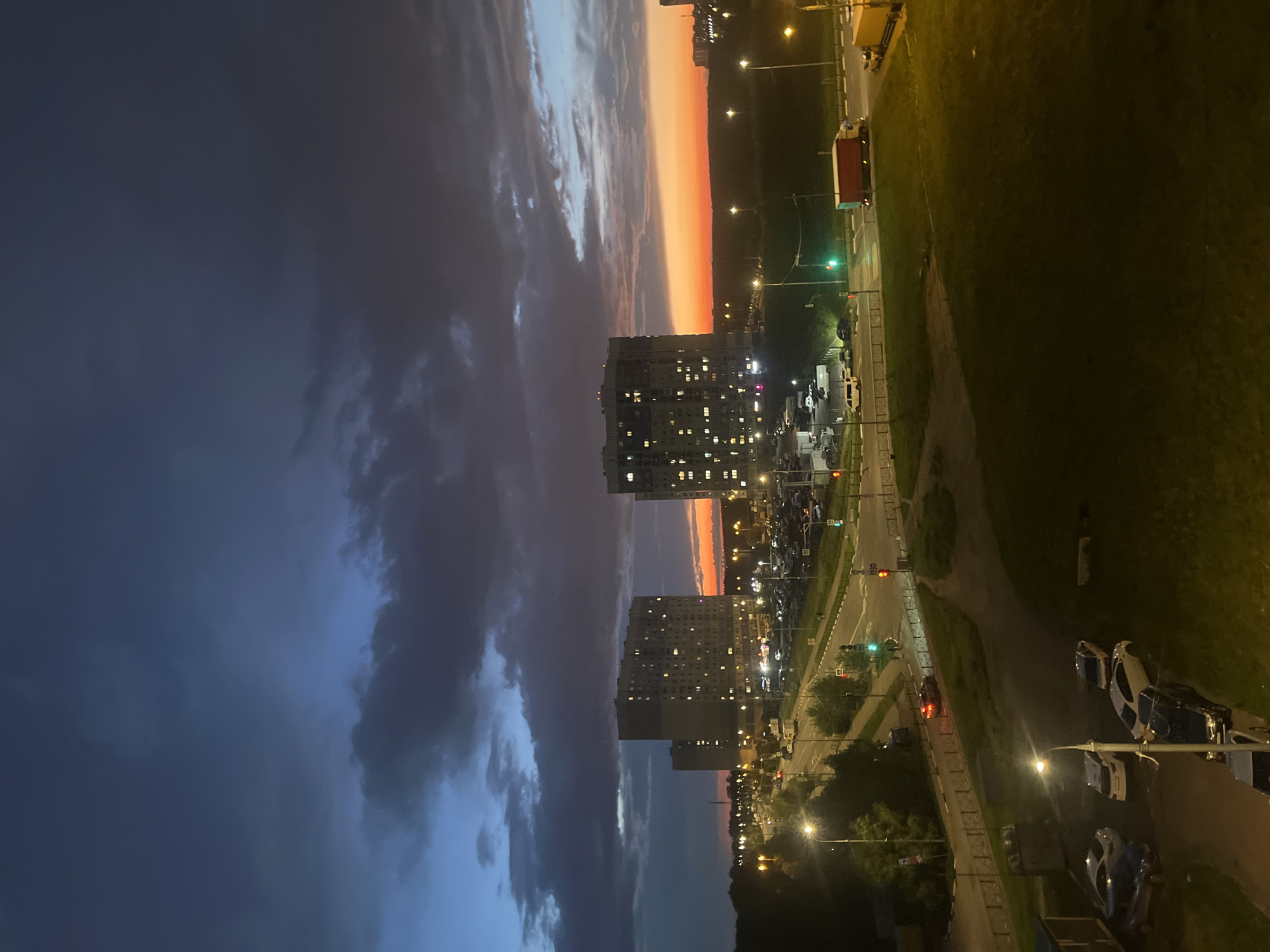 The image depicts a twilight scene in Domodedovo, Russia. The sky is a dramatic canvas of dark, stormy clouds on the left transitioning to vibrant hues of orange and pink at the horizon on the right. Two tall apartment buildings with illuminated windows dominate the mid-ground, suggesting an urban residential area. In the foreground and along the right side of the image, a road with traffic lights and parked cars is visible, with a green grassy area bordering it. The presence of streetlights and car headlights indicates that it is nighttime or the very late evening. The overall mood is serene yet slightly dramatic due to the contrasting sky and the urban environment. There is no visible text in the image.