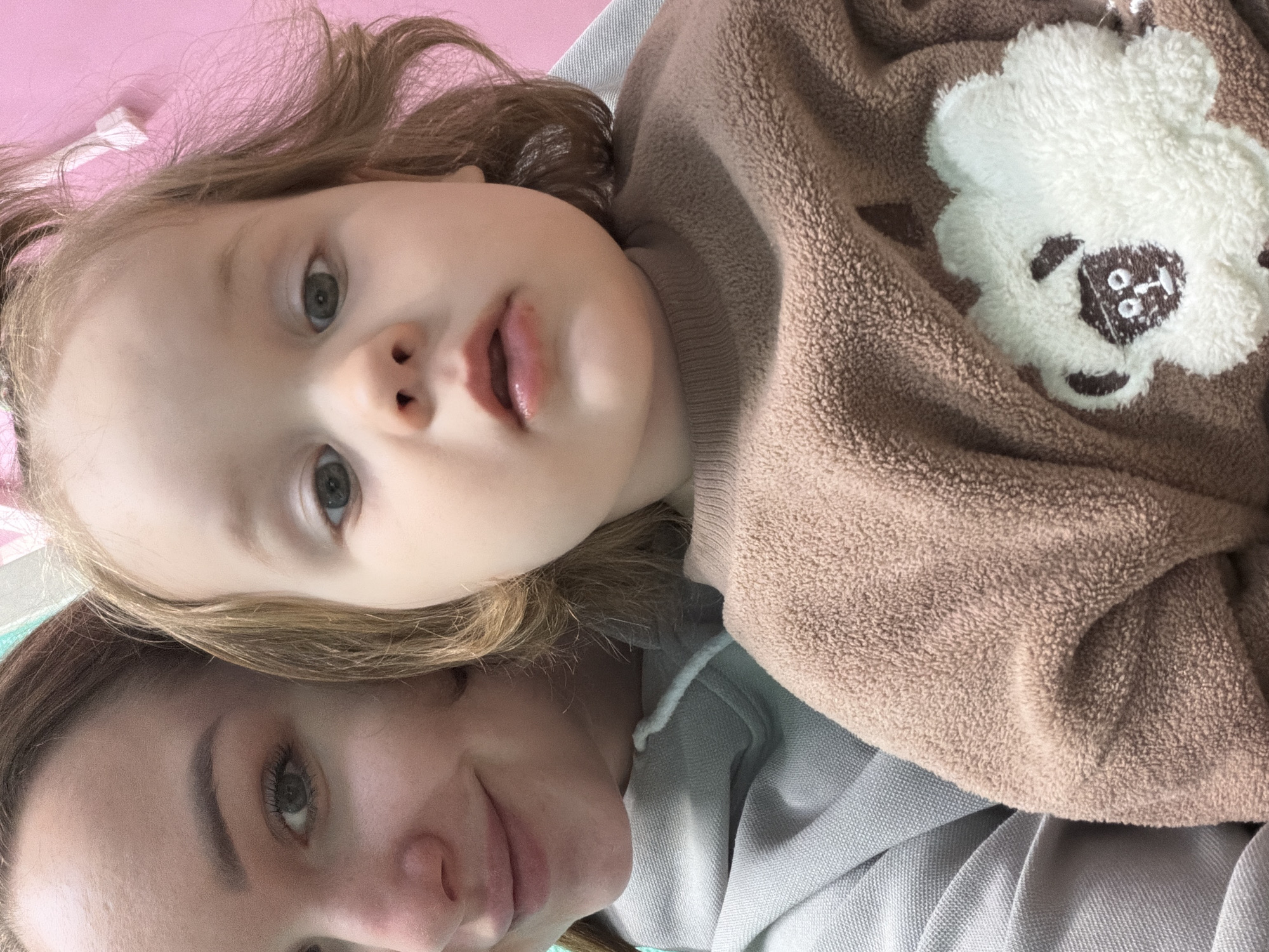 A young child with light hair and blue eyes lies on their back, facing the camera. The child has their mouth slightly open. A woman with brown hair and green eyes is positioned below the child, also facing the camera and smiling slightly. The child is wearing a brown, textured sweatshirt with a white bear applique on the chest. The woman is wearing a light blue hoodie. The background is a pale pink surface. The location information is not discernible from the image.
