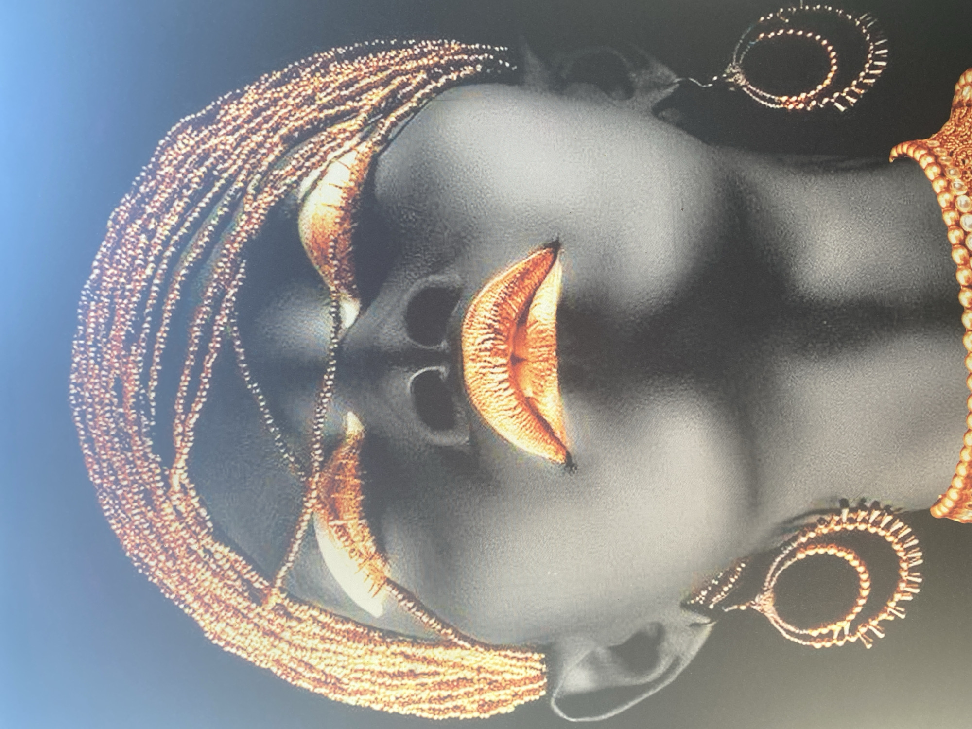 The image shows a close-up, low-angle, portrait of a person adorned with elaborate gold beadwork and makeup. The person's face is painted a dark grey, with striking gold eyeshadow and lipstick. A detailed gold beaded headdress encircles the head, with strands draping across the forehead. Large, circular gold beaded earrings hang from the ears, and a matching beaded necklace is visible at the bottom of the frame. The background is a dark, gradient blue, transitioning to black at the bottom. The image is a studio portrait and does not depict any specific location in Blagoevgrad, Bulgaria.