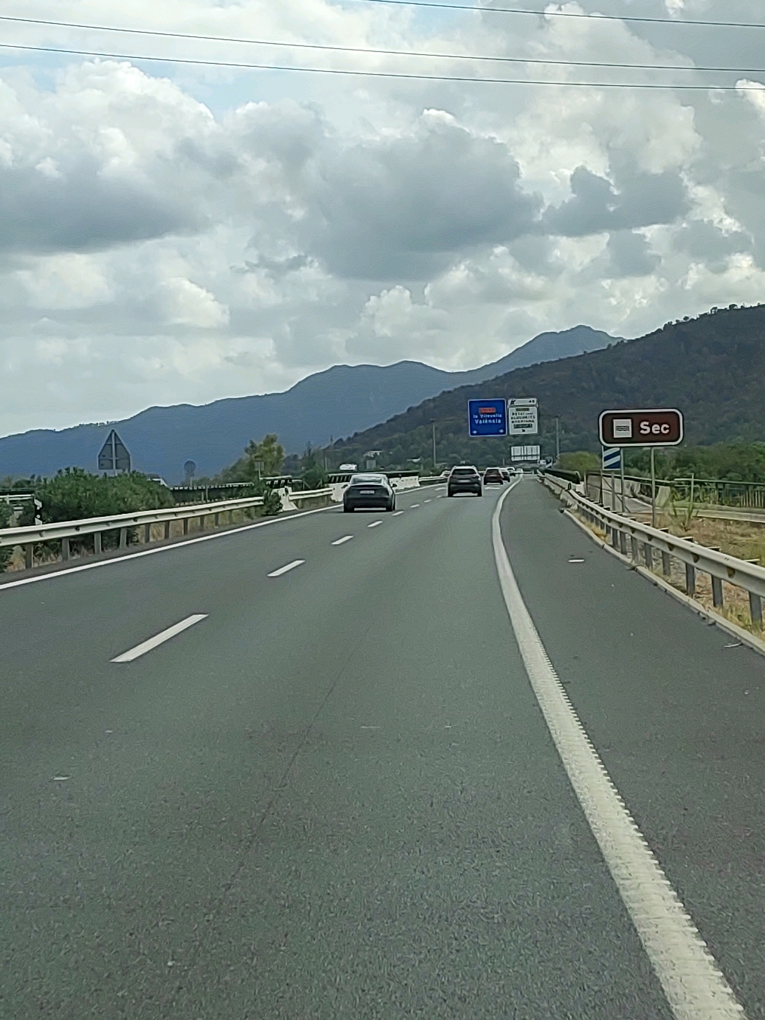 The image depicts a highway scene in the vicinity of Betxí, Spain, under a cloudy sky. The perspective is from a vehicle moving forward, showing a multi-lane asphalt road with clear white lane markings. Guardrails line the sides of the road, with vegetation and hills visible in the background. Several vehicles are traveling on the highway, including a dark sedan and an SUV in the foreground. Road signs are present, including a sign with the text "Sec" in a brown rectangular shape with a wave graphic above it, and other blue signs indicating directions to "La Vilavella" and "València." The overall environment suggests a rural or semi-rural highway setting.
