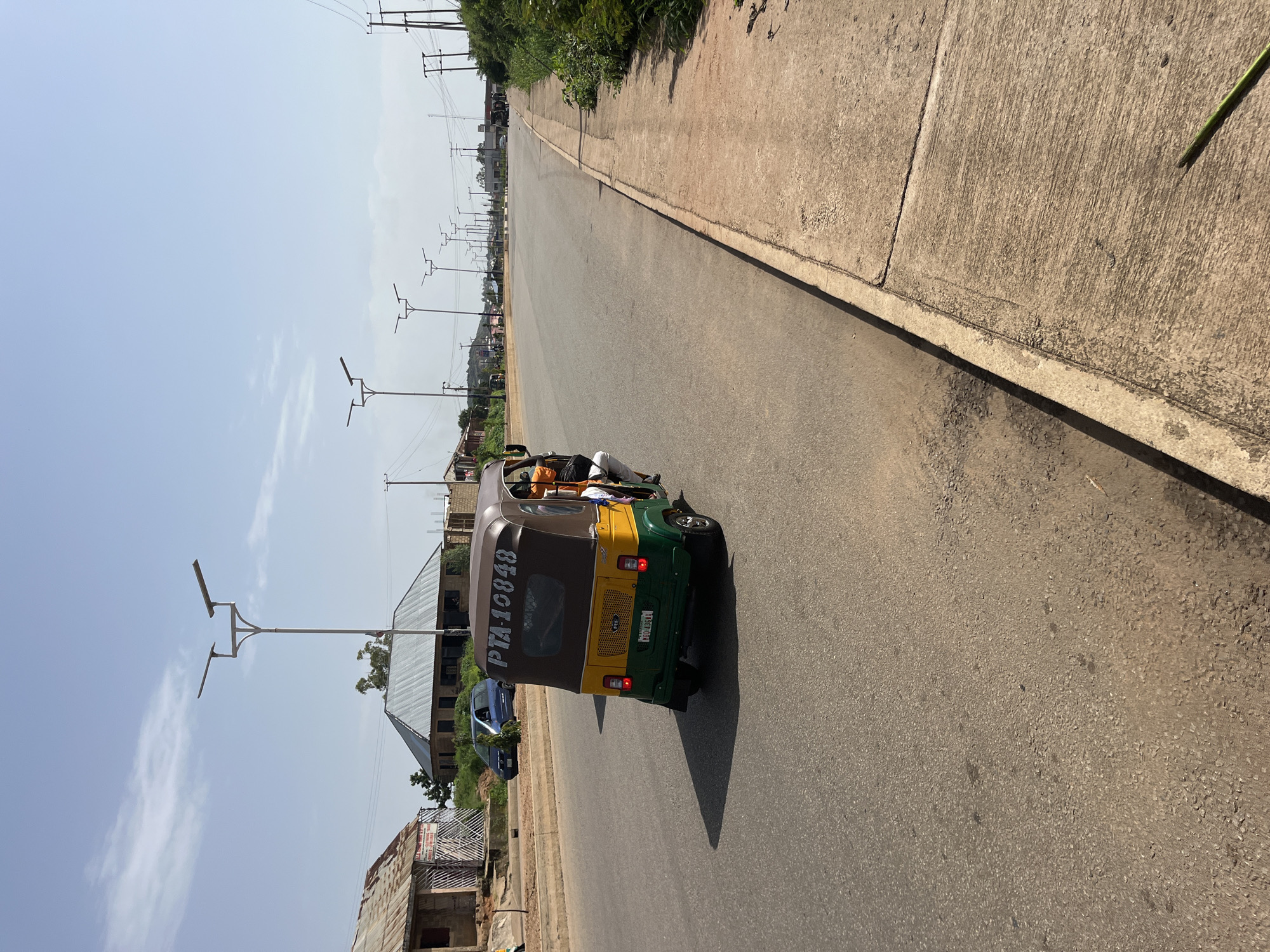 In Bukuru, Nigeria, a three-wheeled vehicle, a Bajaj auto rickshaw, is captured in motion on an asphalt road under a clear sky. The auto rickshaw, painted yellow and green with a brown canopy, displays the registration number "PTA 10848" and a license plate "MGU 436XA". The vehicle is carrying at least one passenger visible in the back wearing an orange shirt. The road is lined with concrete sidewalks and street lights are visible in the distance, suggesting an urban or semi-urban setting. Buildings with corrugated metal roofs are situated along the side of the road. The scene appears to be during daylight hours.