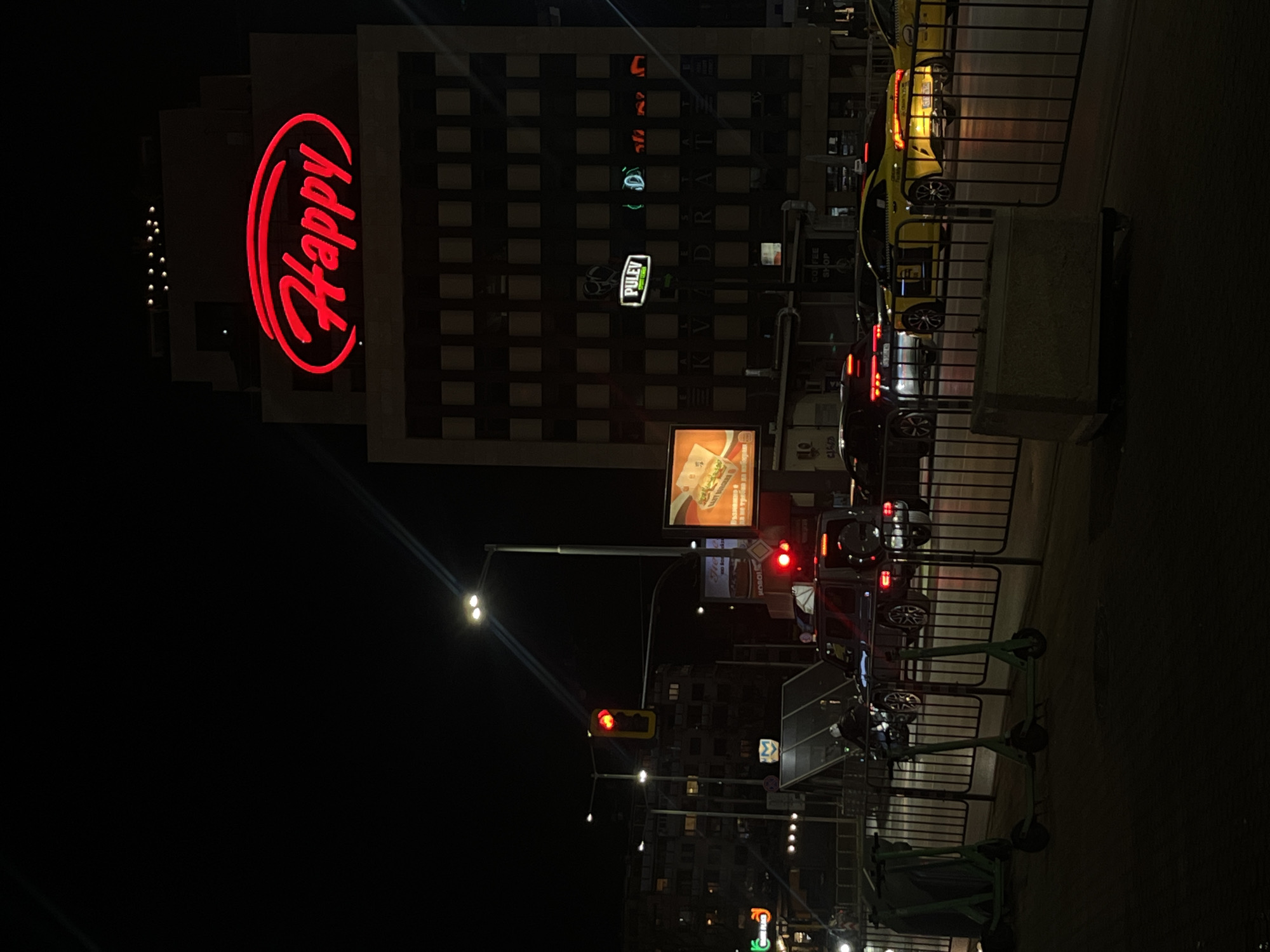 The image captures a night scene in Sofia, Bulgaria, showcasing a brightly lit "Happy" restaurant sign on a building. A mixed-use building is situated to the right of the restaurant. Street-level elements include traffic signals, street lights, parked cars (a yellow one and a dark one), scooters, and fencing that separates pedestrians from the road. Several advertisements appear on buildings. The scene suggests an urban environment with restaurants, traffic, and street furniture.