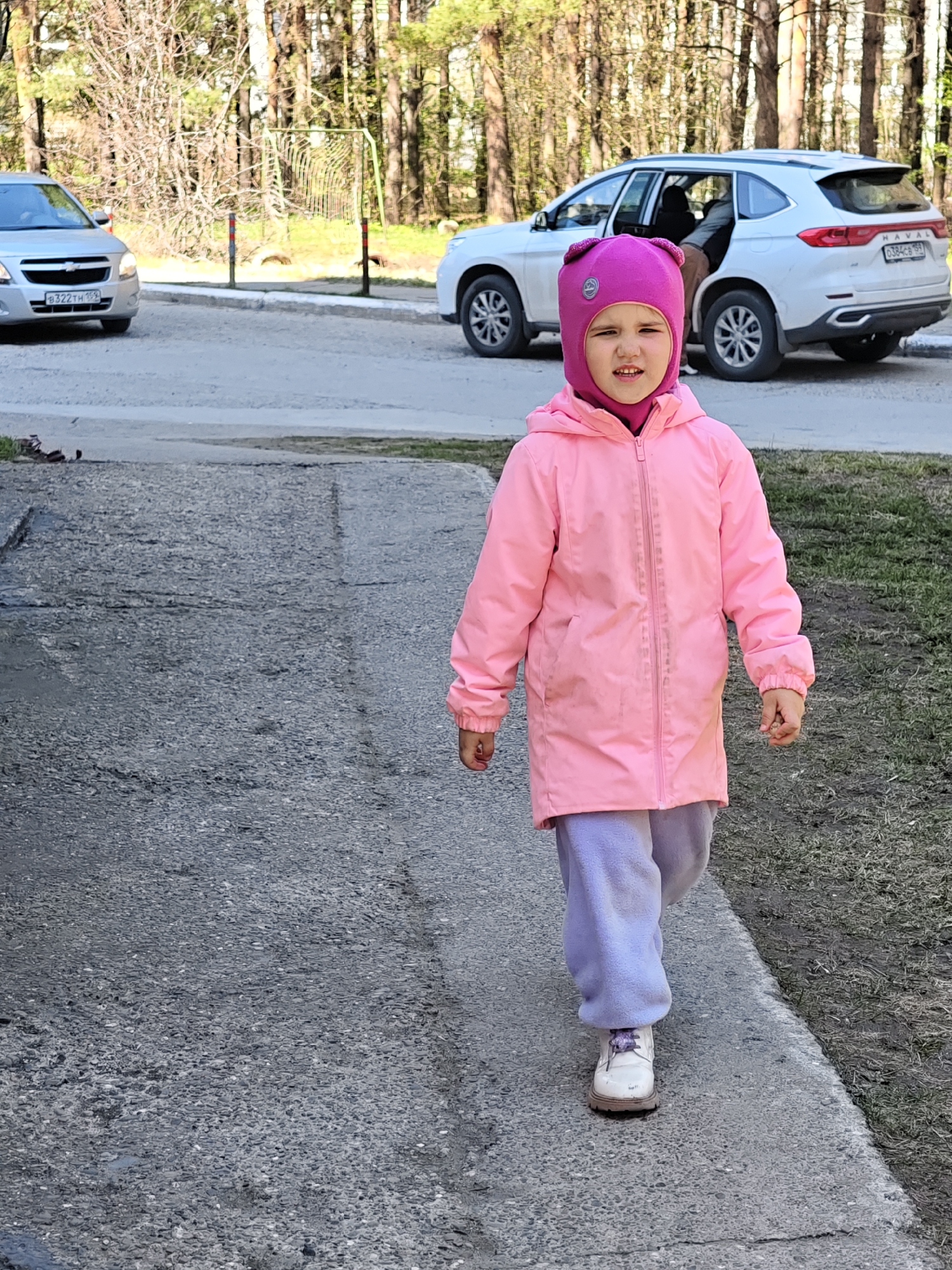 A young girl is walking on a sidewalk in what appears to be a residential area in the city of Chaikovsky, Russia. She is wearing a pink hooded jacket, lavender sweatpants, a pink hat, and white shoes. In the background, there are trees, grass, two parked cars, and a metal soccer goal. The license plate on the silver car on the left is "B322TH 159". The license plate on the white SUV is "O384CE 59". The sidewalk and road are made of asphalt.