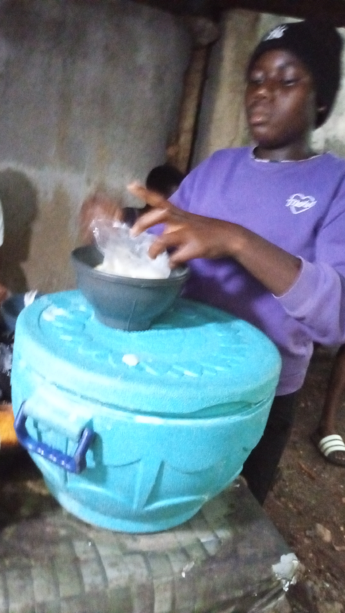 A young woman wearing a black beanie and a purple long-sleeved sweatshirt stands next to a large, blue, insulated container in what appears to be a dimly lit interior or outdoor area. She is holding a plastic bag over a black bowl that sits on top of the container, as if transferring something into the bowl. The background shows parts of a concrete wall and the legs of other individuals. The setting is suggested to be Adankolo, Nigeria, based on provided context. The surface beneath the container is covered with what seems to be a tarp or mat.