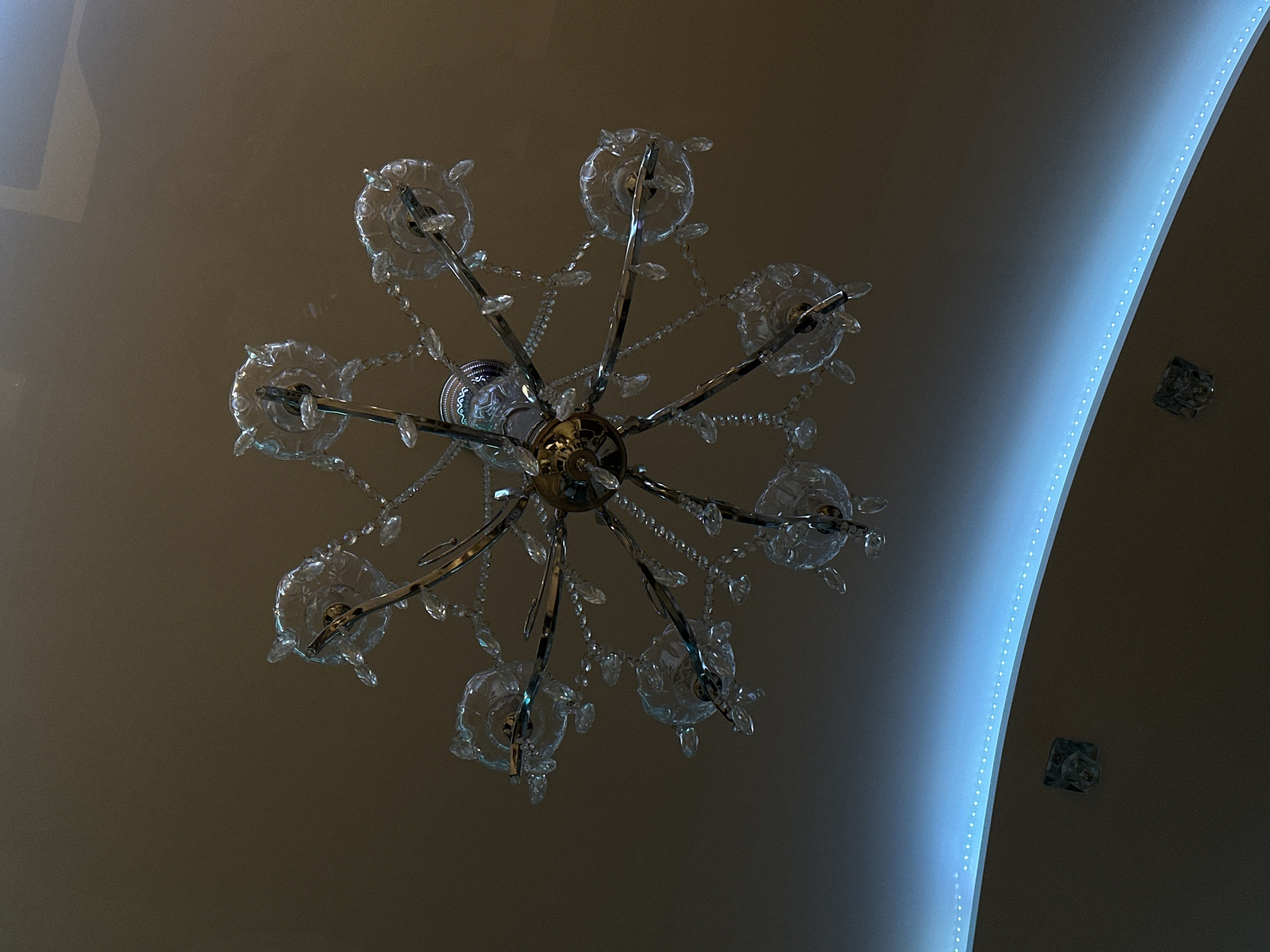 The image shows a chandelier hanging from a ceiling. The chandelier has a central metallic structure with multiple arms extending outwards. Each arm holds a glass or crystal element. There are additional crystal chains hanging between the arms. The ceiling is a light brown color with a curved, recessed section that has a blue light strip running along its edge. There are also two square, glass-like objects mounted on the ceiling near the blue light. While the location in the city of Khimki, Russia, is mentioned, there is no additional information in the image that relates to this location.