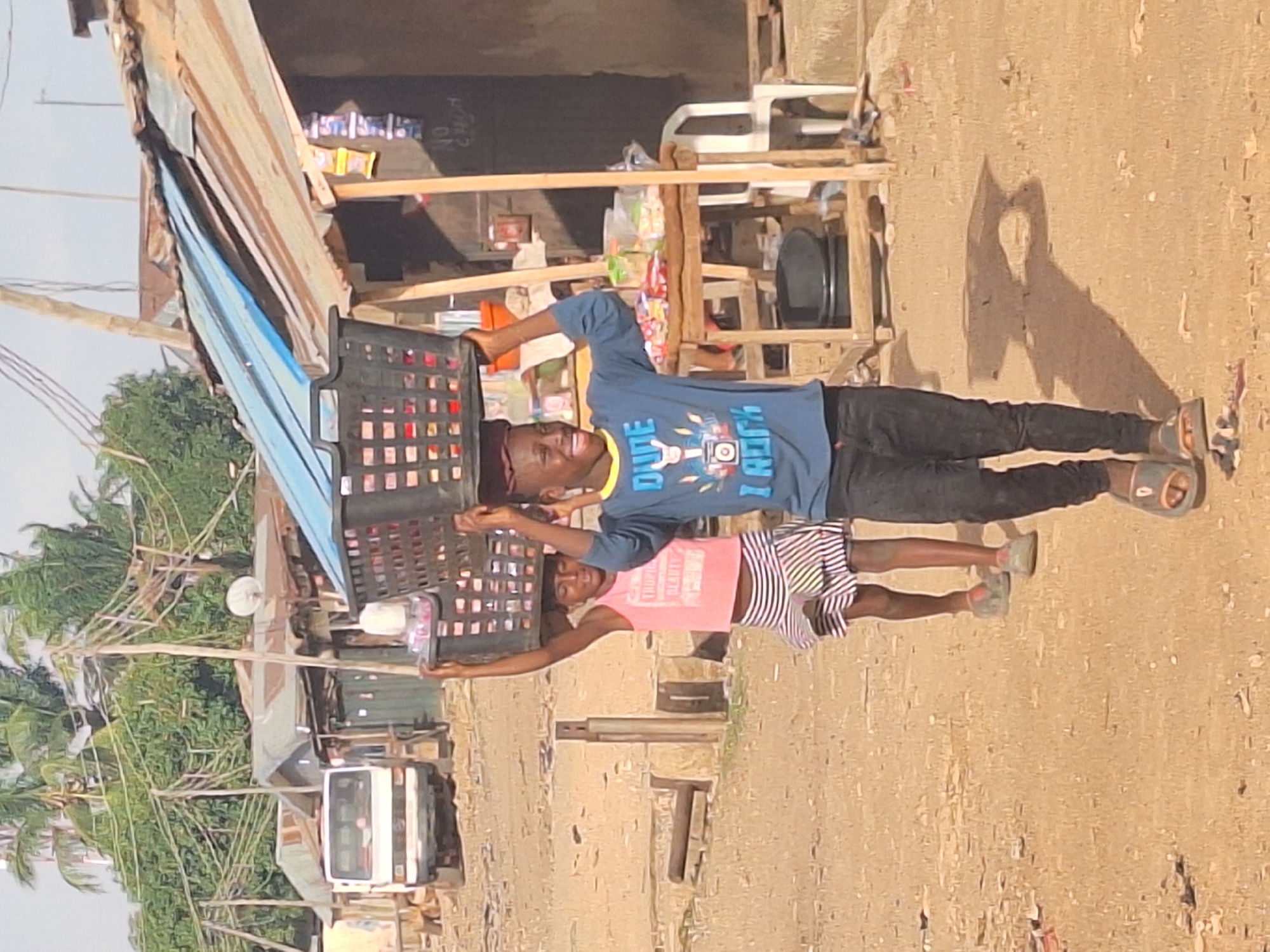 Two children are carrying a black plastic crate between them outdoors in Sabon Tasha, Nigeria. The child on the top wears a blue t-shirt, black pants, and sandals. The child below wears a pink t-shirt and striped shorts and green shoes. The ground is dirt, and there is a building to the left with a metal roof. There is a white van, palm trees, and a small shop in the background. The sky is light. The image is rotated to the right.