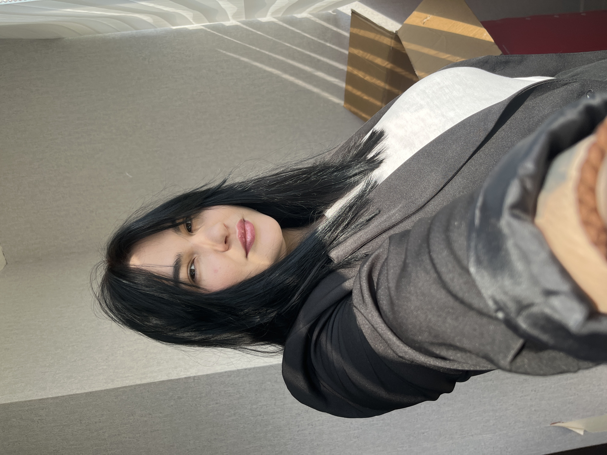 A woman with long, straight dark hair, fair skin, and dark eyes is depicted in a close-up, slightly tilted portrait. She has a neutral to slightly pleasant expression, with her lips tinted pink. She is wearing a dark-colored, possibly black or very dark grey, hooded jacket or top over a white inner garment. The image is taken indoors in Chaikovsky, Russia.
The background consists of a plain, light-colored wall or surface. Strong, distinct horizontal shadows from what appear to be window blinds are cast across the upper part of the background and onto a partially visible brown cardboard box located to the upper right of the woman. This bright, directional lighting indicates that the photograph was taken during the daytime, likely when the sun is low in the sky, such as morning or late afternoon. No specific activities are ongoing other than the pose for the photo. There is no visible text in the image.