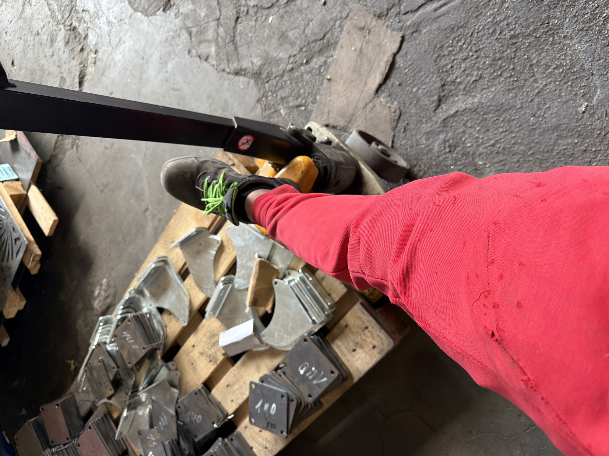 Observed is an overhead shot in what appears to be an industrial setting. A leg clad in red fabric is positioned over a pallet filled with metal components, of which some are stamped with numerals. One foot is shod in a gray shoe with lime green laces. A piece of machinery with a black arm extends into the view. The ground is concrete and chipped. Location is Unknown, Unknown.
