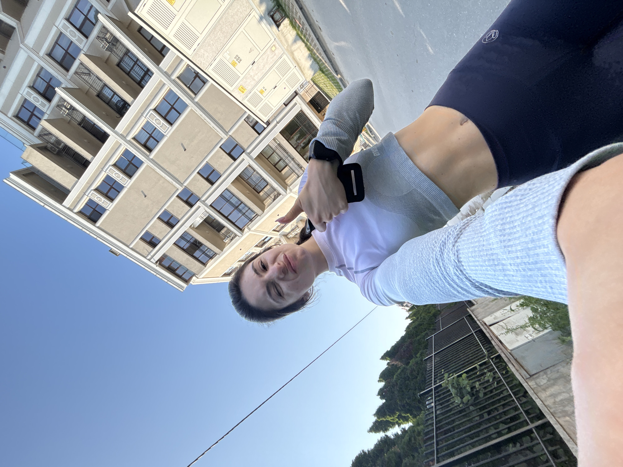 A rotated outdoor selfie captures a young woman giving a thumbs-up gesture. The image is oriented 90 degrees clockwise, with the woman positioned centrally, looking up at the camera.
The woman has dark hair pulled back and a cheerful expression, smiling. She wears a light grey long-sleeved athletic top, a white short-sleeved top underneath, and dark blue or black athletic leggings, which reveal her midriff and navel. A black smartwatch is on her left wrist, and she holds a black rectangular object, possibly a phone, in the same hand while extending her thumb upwards. A small, indistinct white logo is visible on her left upper thigh.
In the background, a multi-story residential building dominates the left side of the frame (when upright). It features a light-colored (beige or off-white) facade with numerous dark-framed windows and balconies with black railings. Decorative architectural elements surround the windows. Several external utility boxes with yellow, unreadable labels are mounted on the building.
The right side of the image (when upright) shows a clear blue sky, indicating bright, sunny daytime weather. A thin black cable stretches horizontally across the lower middle of the sky. Below the cable, green trees and dense foliage are visible, alongside a dark metal fence or grate structure. The ground beneath the woman's legs appears to be paved with asphalt or concrete.
The bright natural light suggests it is daytime. The overall scene conveys a positive and active mood. The architecture of the building is consistent with an urban or suburban setting, aligning with the provided context of Varna, Bulgaria.