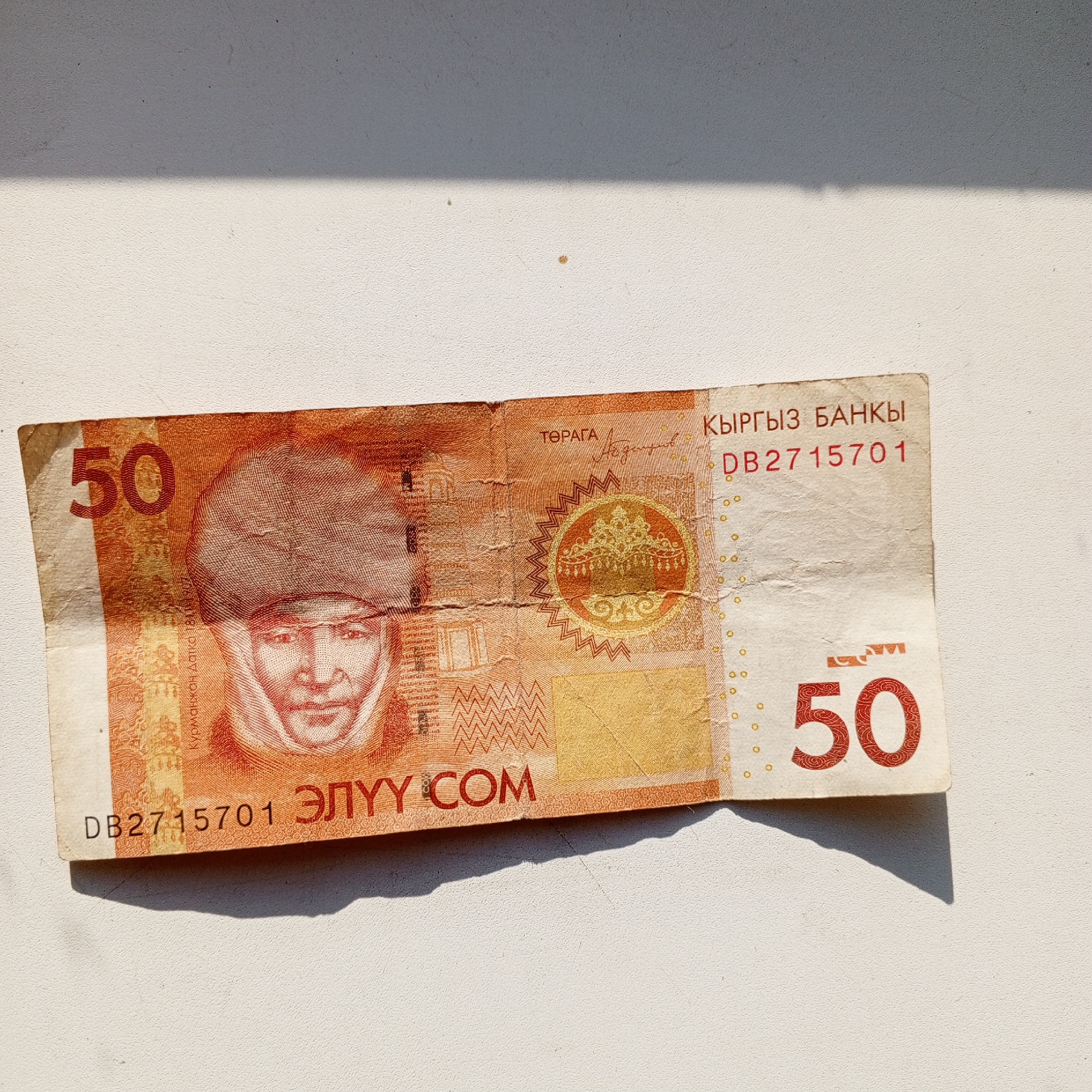 The image shows a close-up of a wrinkled, orange and white banknote. The banknote has a portrait of a man with a hat on the left side, and the number "50" is prominently displayed in red on both the left and right sides. Text in Cyrillic script is visible, including "КЫРГЫЗ БАНКЫ" (Kyrgyz Bank) and a serial number "DB2715701". There is also a circular emblem with intricate patterns. The banknote is lying on a textured, light-colored surface, with a shadow cast across the top portion of the image. The image is taken from a top-down perspective. No specific location in Sayanogorsk, Russia, can be determined from the image itself.