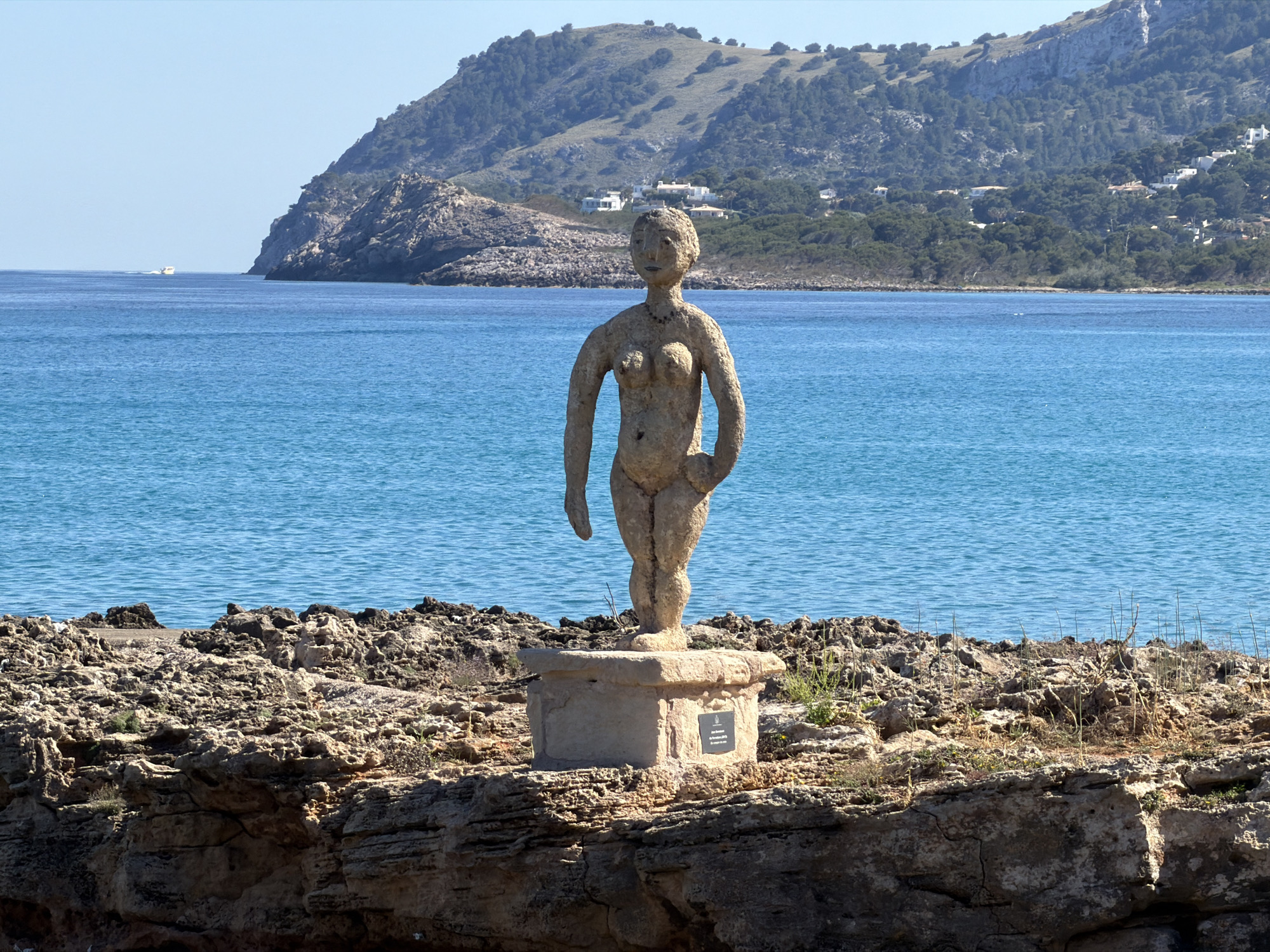 A full shot shows a nude sculpture of a woman standing on a plinth in front of a bay. The sculpture is made of a light-colored material that appears to be stone or concrete. The woman is standing with her left arm at her side and her right hand on her hip. The plinth is a simple square shape, and there is a small plaque on the front of it. The bay is calm and blue. In the distance, there is a hill covered with trees. The location is Capdepera, Spain.