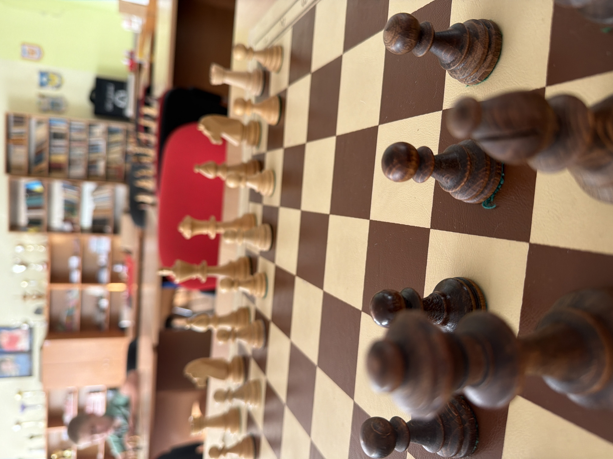 The image shows a close-up of a wooden chessboard with chess pieces. The board is set up for a game, with both white and dark-colored pieces arranged in their starting positions. The perspective is from an elevated angle, looking down and across the board. The light-colored squares are a tan or beige color, and the dark squares are a deep brown.
On the dark side, several pieces are visible, including pawns and what appear to be a rook or bishop further back. These pieces are made of dark wood and have a classic Staunton design. On the white side, a line of pawns and a knight are clearly visible, also in a traditional design. The white pieces are made of a lighter wood, possibly boxwood or maple.
In the background, out of focus, a room is visible. There are shelves filled with books and other items, suggesting a library, study, or classroom setting. A red chair is partially visible, and a black bag with a white logo is on one of the shelves. A person's head and shoulders are visible in the lower left corner, appearing to be looking down at the chessboard. The lighting suggests it might be daytime, with natural light contributing to the illumination. There is no visible text that can be clearly discerned. The overall impression is a scene of a chess game in progress within an indoor, possibly academic or recreational, environment.