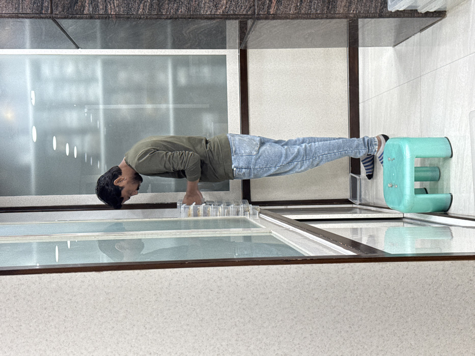 A man is performing a plank exercise, using the edge of a glass-topped counter for hand support and a light blue stool for foot support. He wears a long-sleeved green shirt and blue jeans with striped socks. The background consists of a wall with paneling and a section of a window with light reflections. Six small transparent containers are arranged along the counter's edge, in front of him. The image is rotated 90 degrees clockwise.