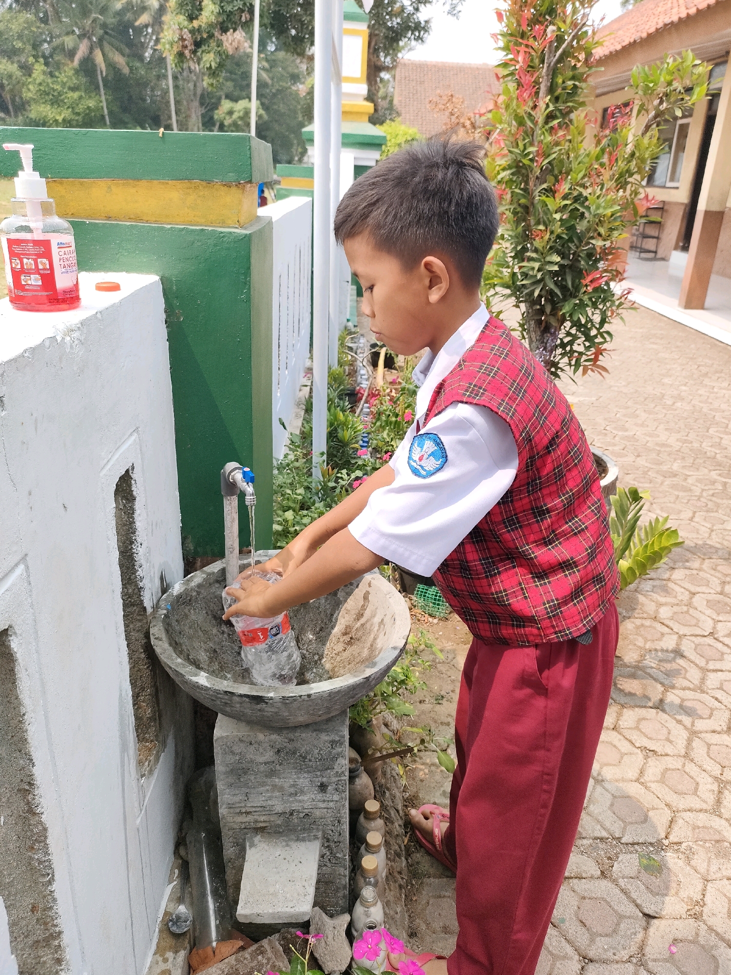 In Bojong, Indonesia, a young boy in a red and white plaid vest and maroon pants washes a plastic water bottle under an outdoor faucet. The faucet, attached to a pipe, drips water into a concrete sink. The sink is positioned below a white wall, which has a green and yellow painted structure above it. To the left of the sink, a bottle of hand sanitizer sits on the white wall. The scene is set on a paved walkway, with lush greenery and trees surrounding the area, and a building with a red tiled roof visible in the background.