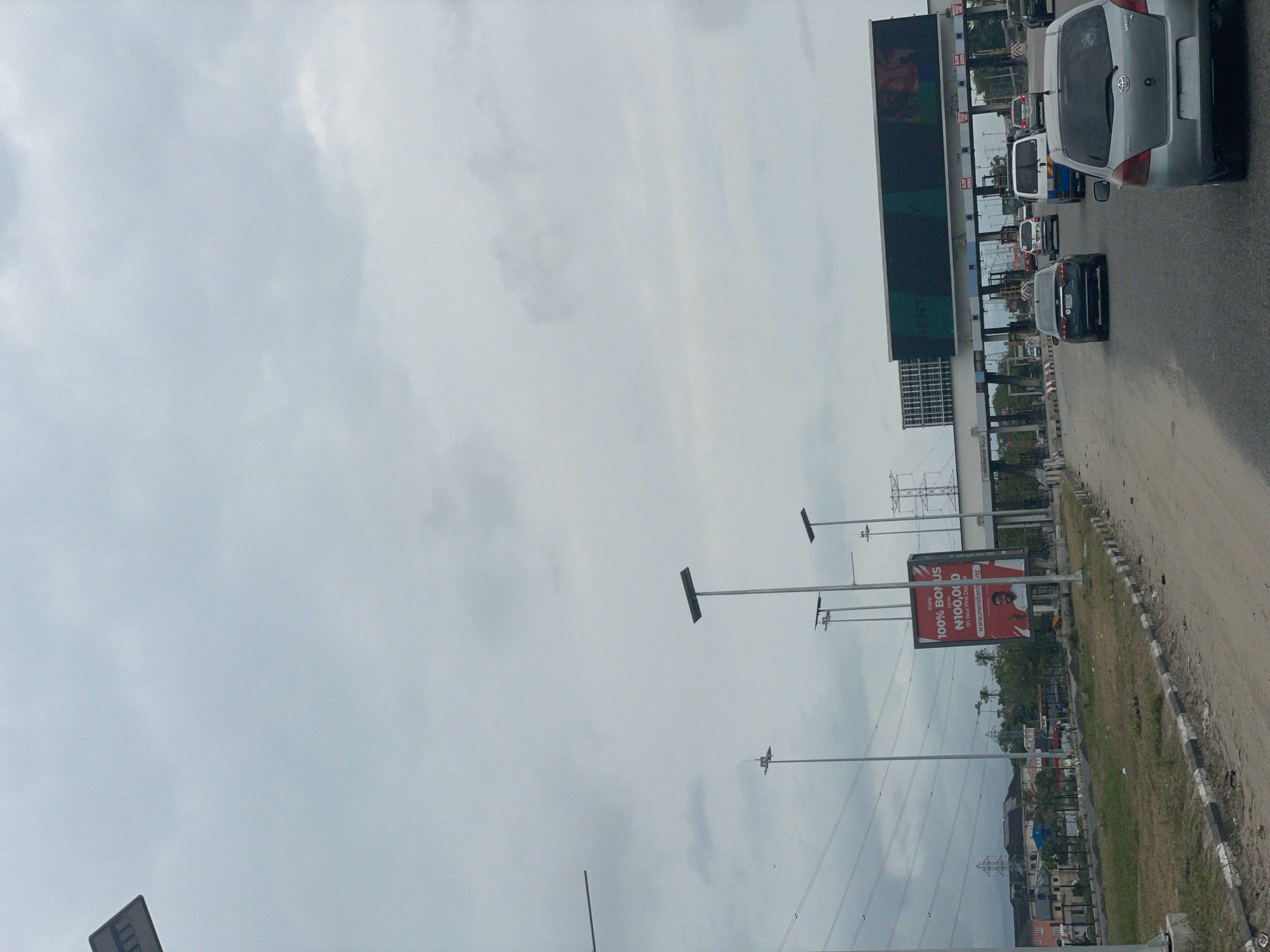 The image depicts a street scene in Lekki, Nigeria, characterized by a busy tollgate. Cars are lined up, waiting to pass through the toll booths. A large electronic billboard displays a commercial advertisement above the toll booths. Adjacent to the roadway, a sign advertises a "100% BONUS" of up to "N100,000" on a first deposit. Utility poles and power lines run parallel to the road. The sky is overcast and cloudy. Buildings and vegetation are visible in the background.